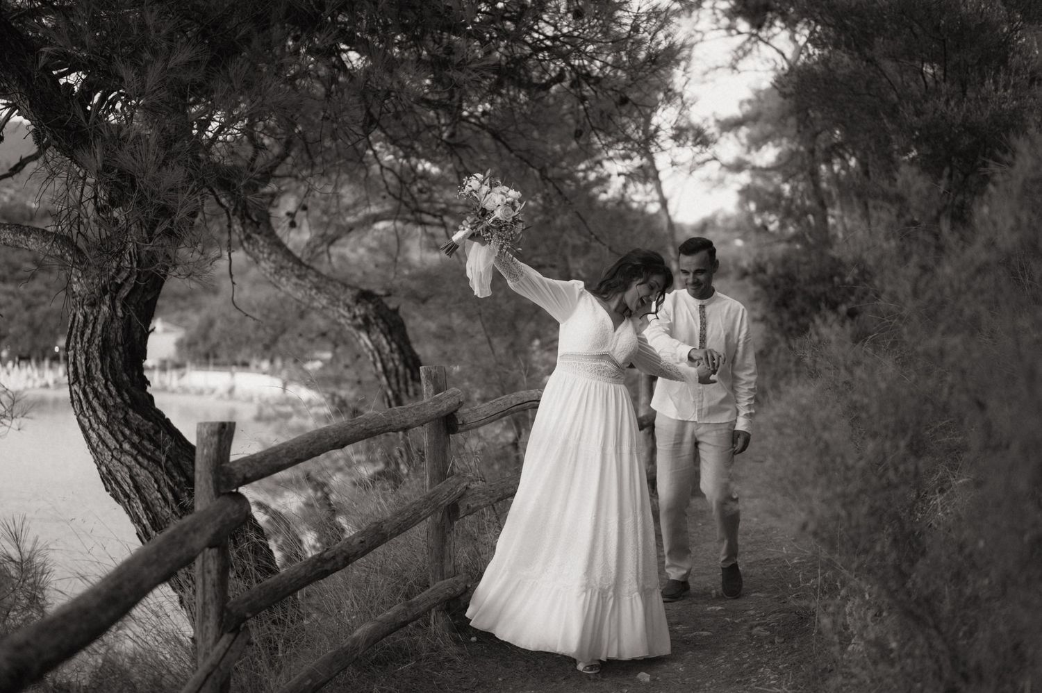 Bride in white gown dancing with groom on a scenic tree-lined path with wooden fence in black and white.
