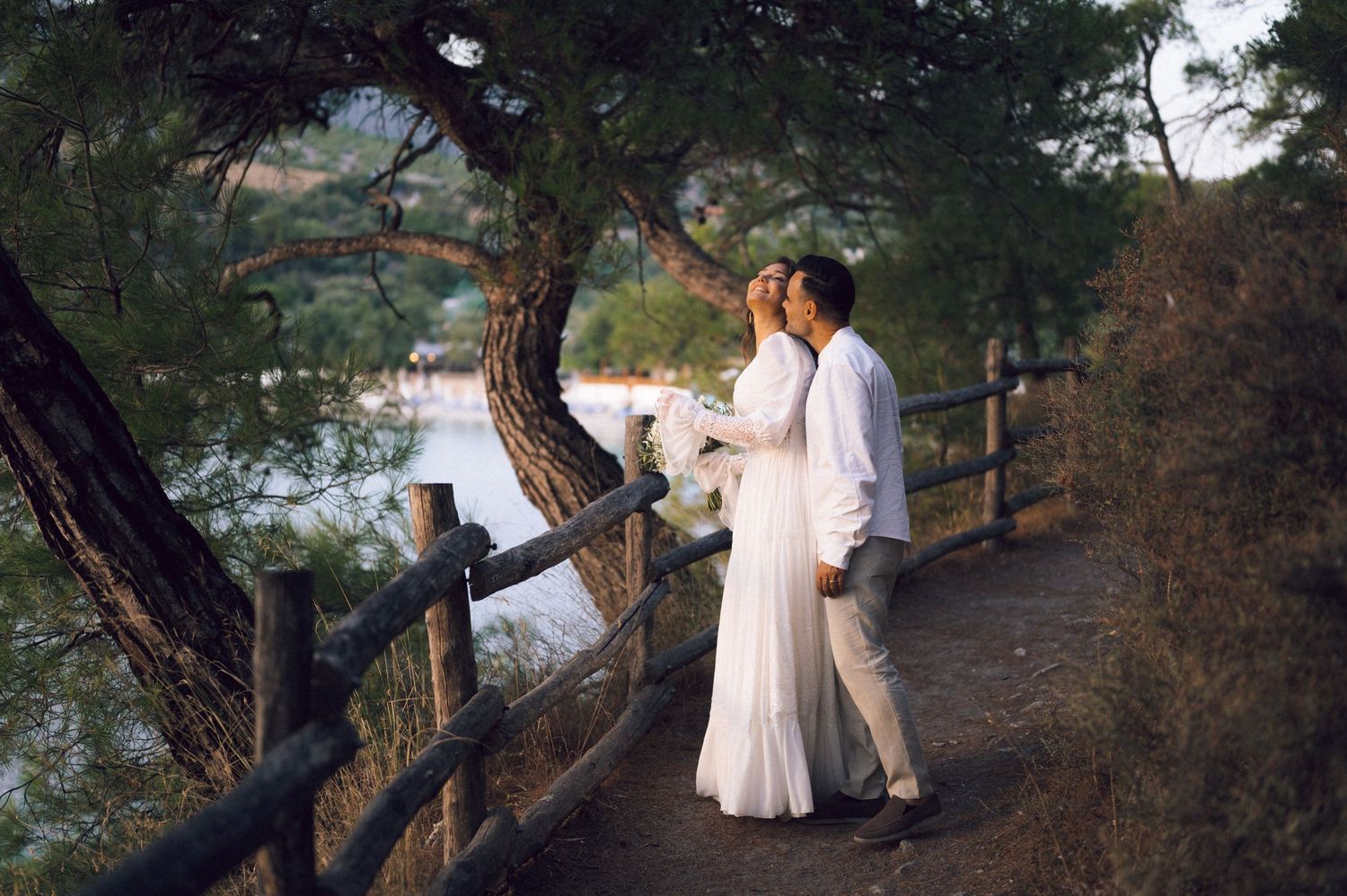 A couple in white attire shares a romantic moment on a scenic lakeside path surrounded by lush green trees.