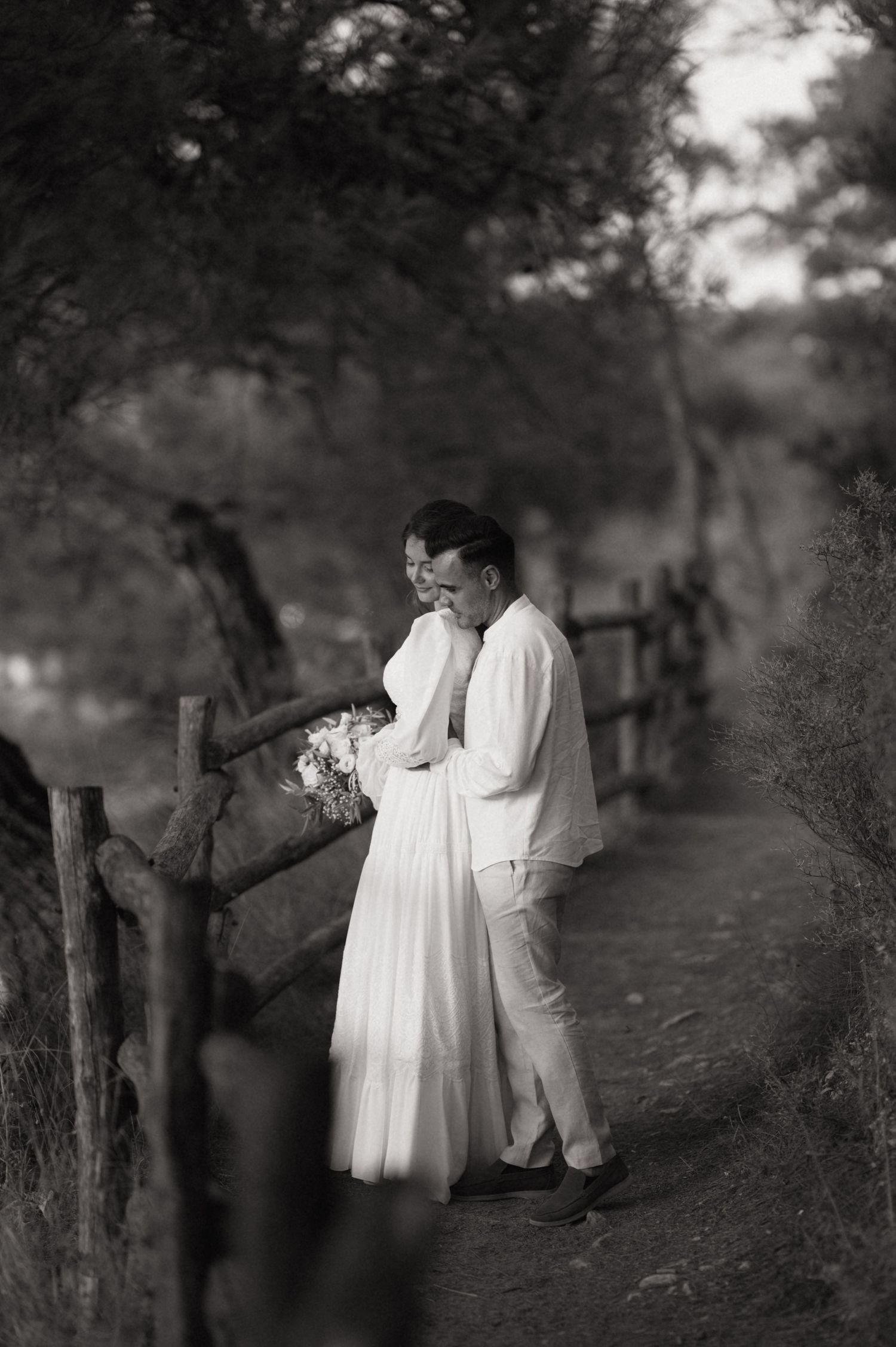Black and white photo of a couple in white attire embracing on a rustic path surrounded by trees.