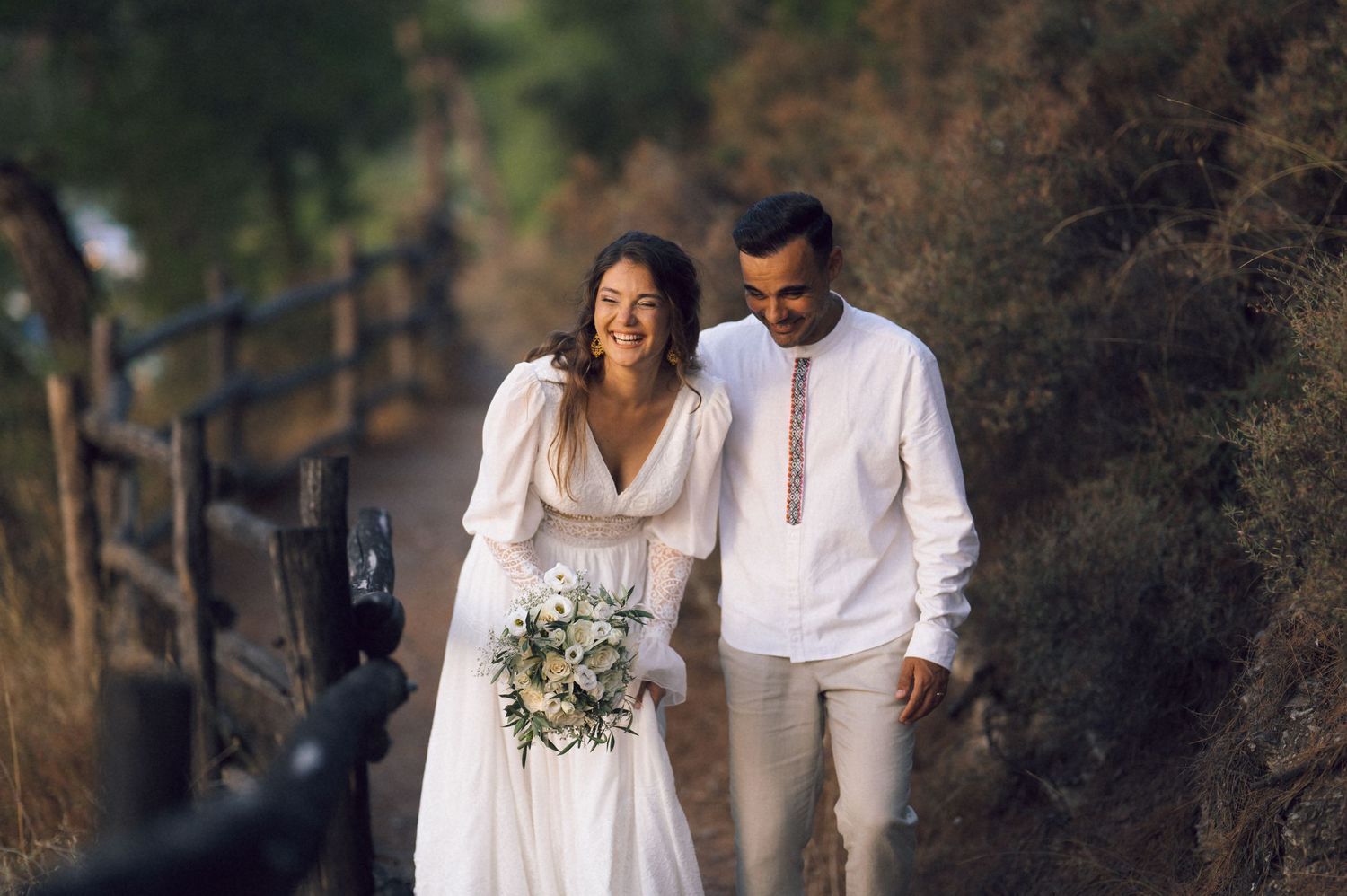 A smiling bride in a white gown holding a bouquet walks with a groom in white on a scenic outdoor path.