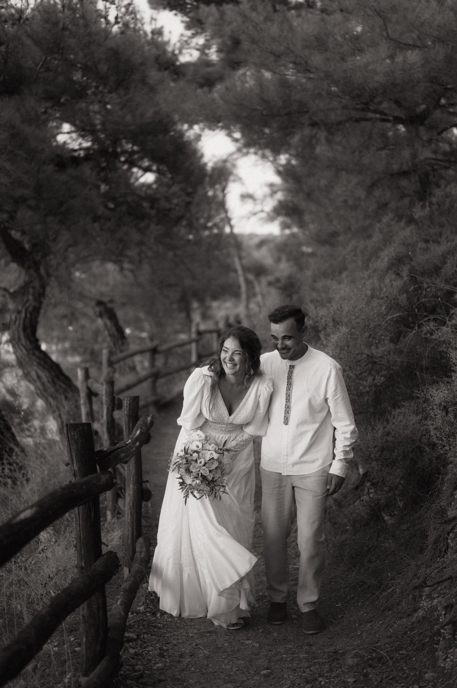 Black and white photo of a smiling bride and groom walking a tree-lined path, bride holding a bouquet.