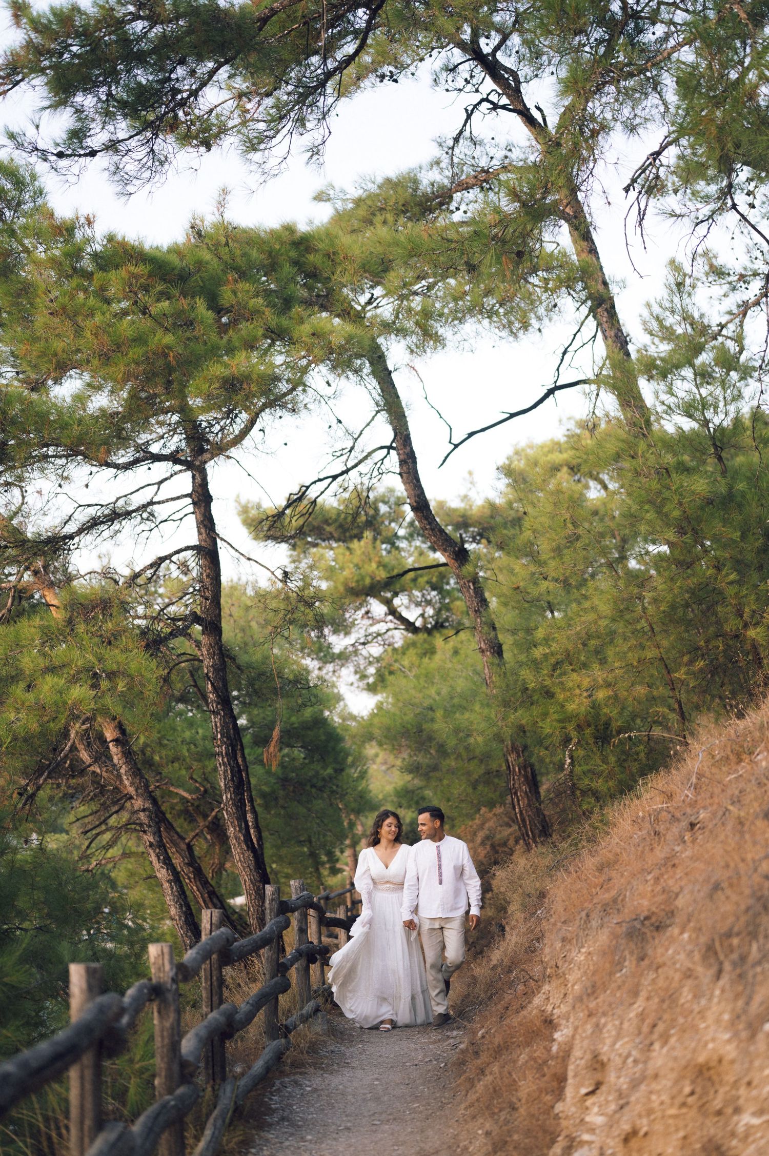 Couple in white attire walking down a scenic forest path with pine trees and wooden fence.