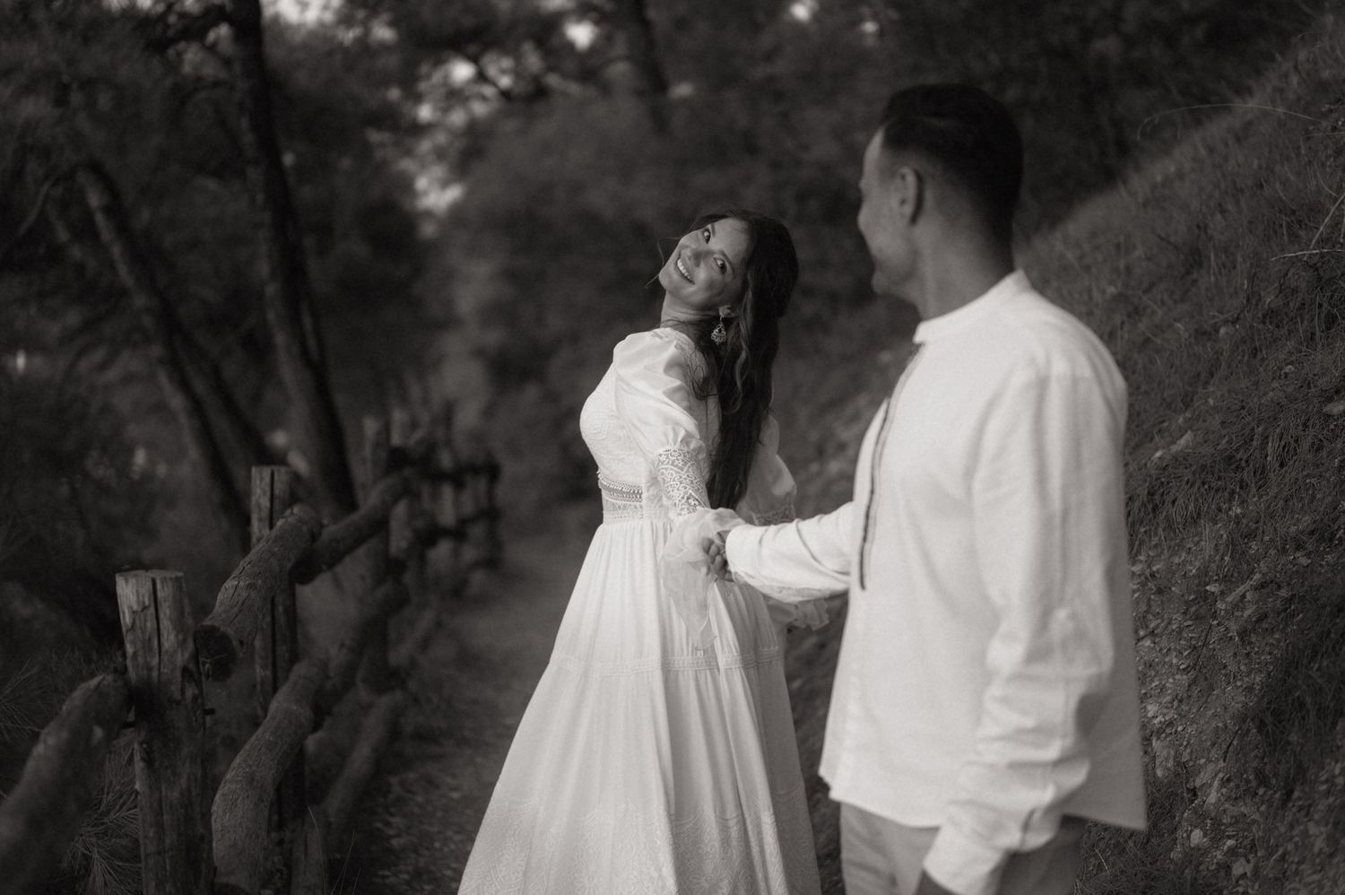 Black and white photo of a couple in white attire sharing a romantic moment on a forest path near a wooden fence.