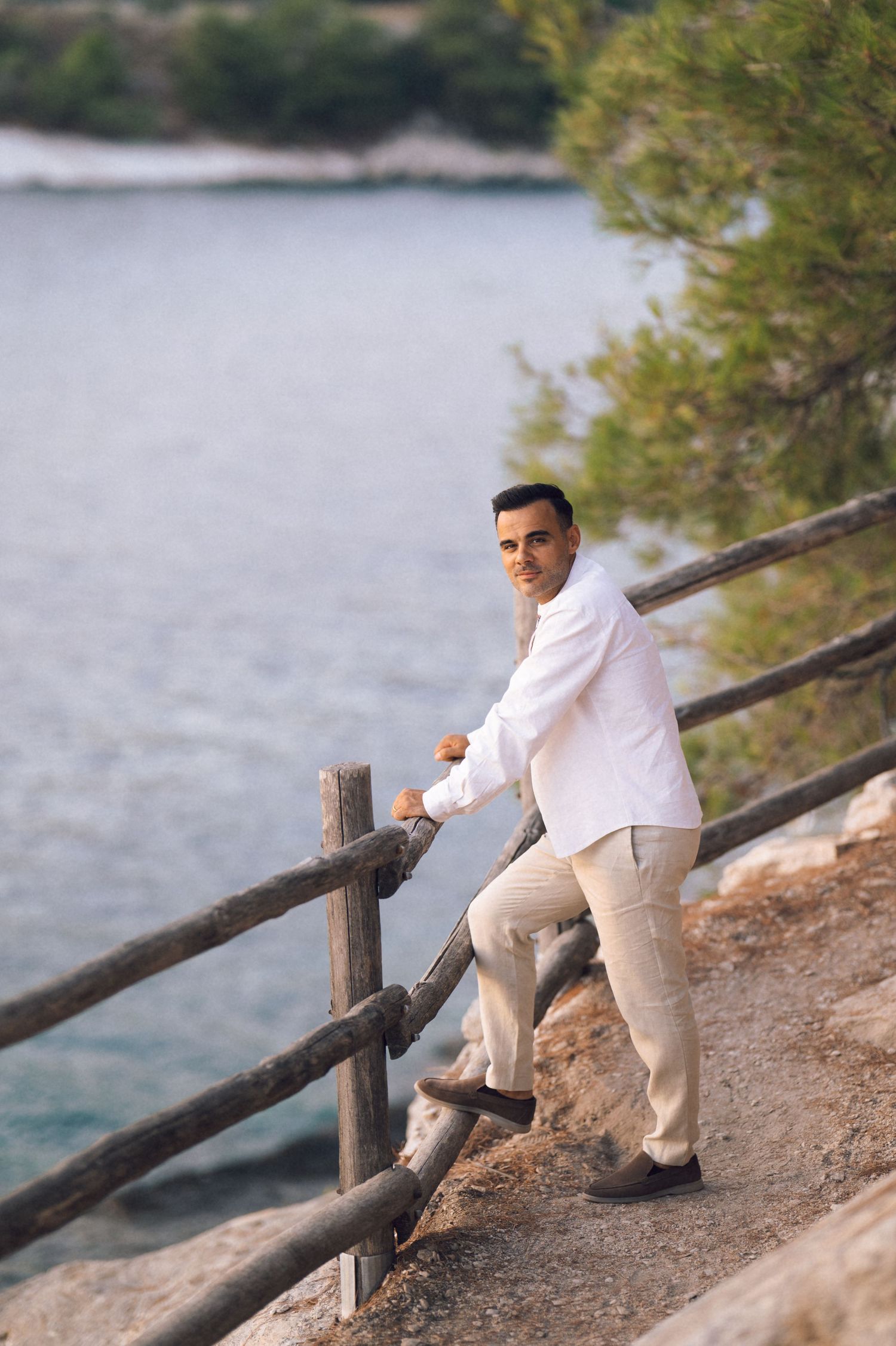 Man in white shirt and beige pants leaning on wooden fence railing by the sea on a rocky coastal path.