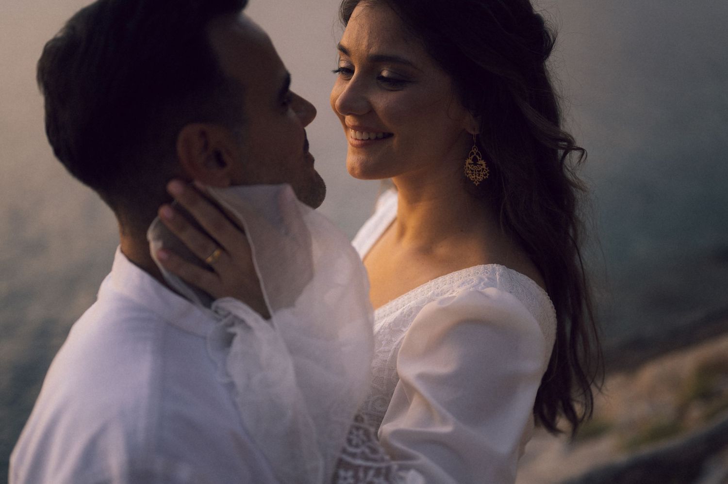 Romantic couple in white outfits sharing an intimate moment outdoors with scenic mountain backdrop.