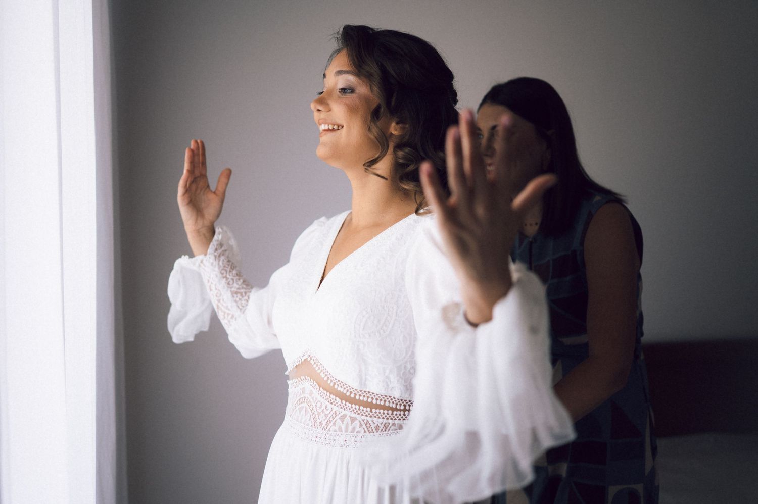 Bride in white lace dress smiling near window while bridesmaid helps her prepare for wedding day.