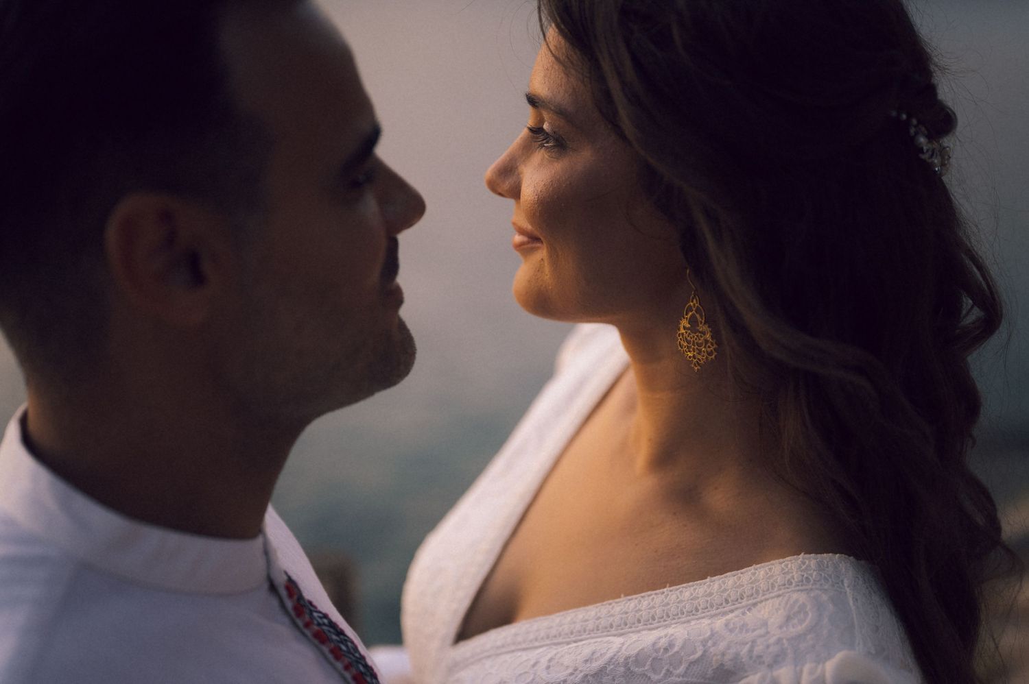 Romantic couple in close embrace, woman smiling in off-shoulder white dress with gold earrings, soft moody lighting.