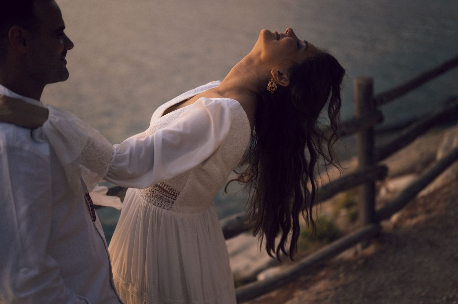 A couple shares a romantic moment near a wooden fence overlooking the ocean at sunset, woman in white dress.