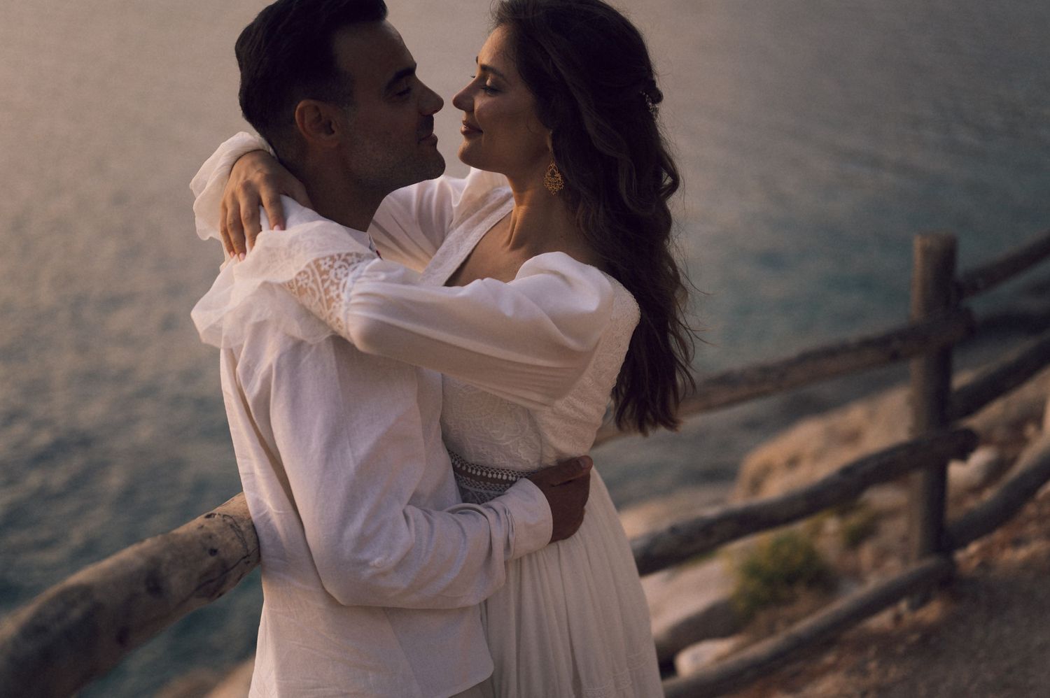 Romantic couple embracing at sunset near ocean cliff, woman in white dress, golden hour light.
