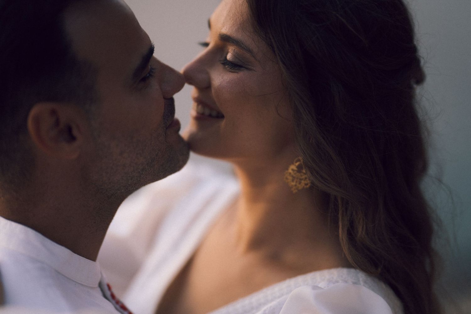 A romantic couple sharing an intimate moment, the woman wearing gold earrings and a white outfit, smiling softly.