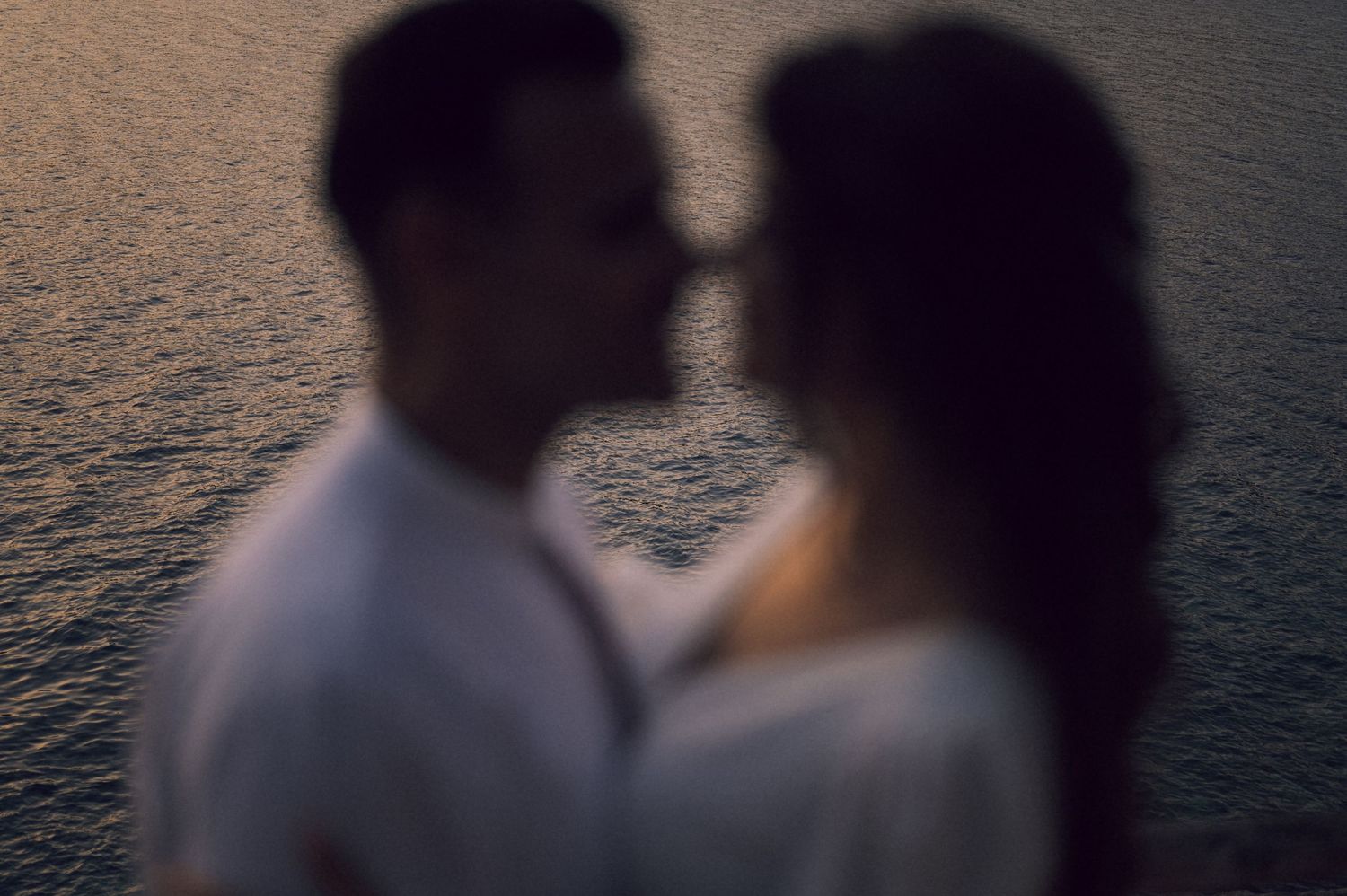 Romantic couple sharing an intimate moment near shimmering water at dusk in a soft focus portrait.