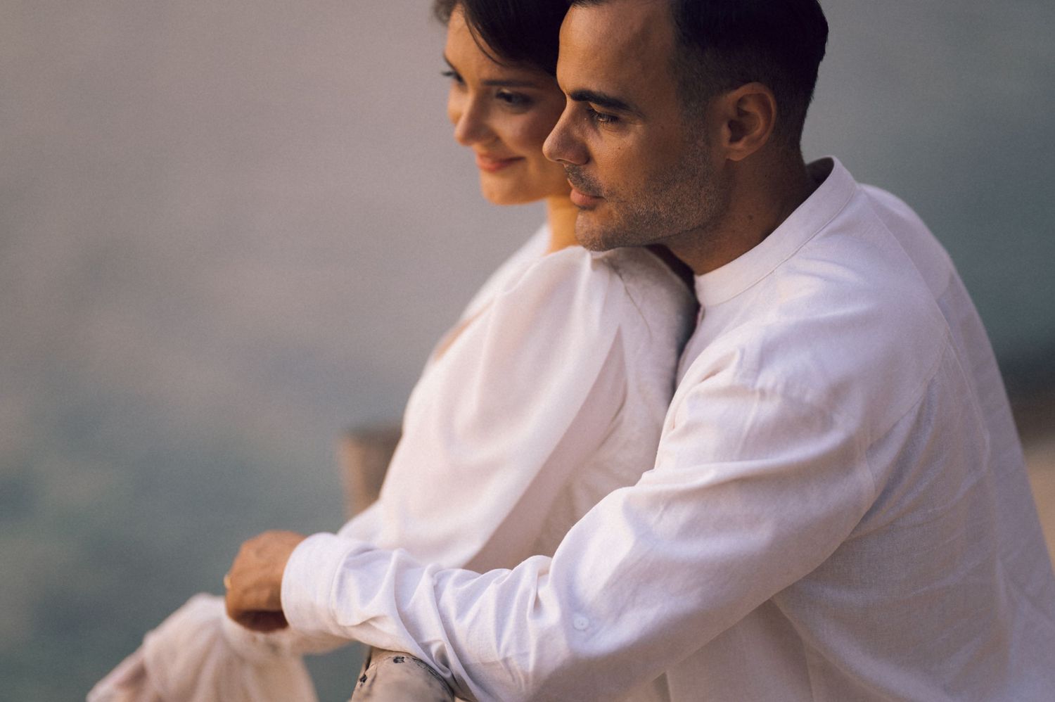 A couple in white outfits sitting together outdoors, sharing a tender moment with a soft blurred background.