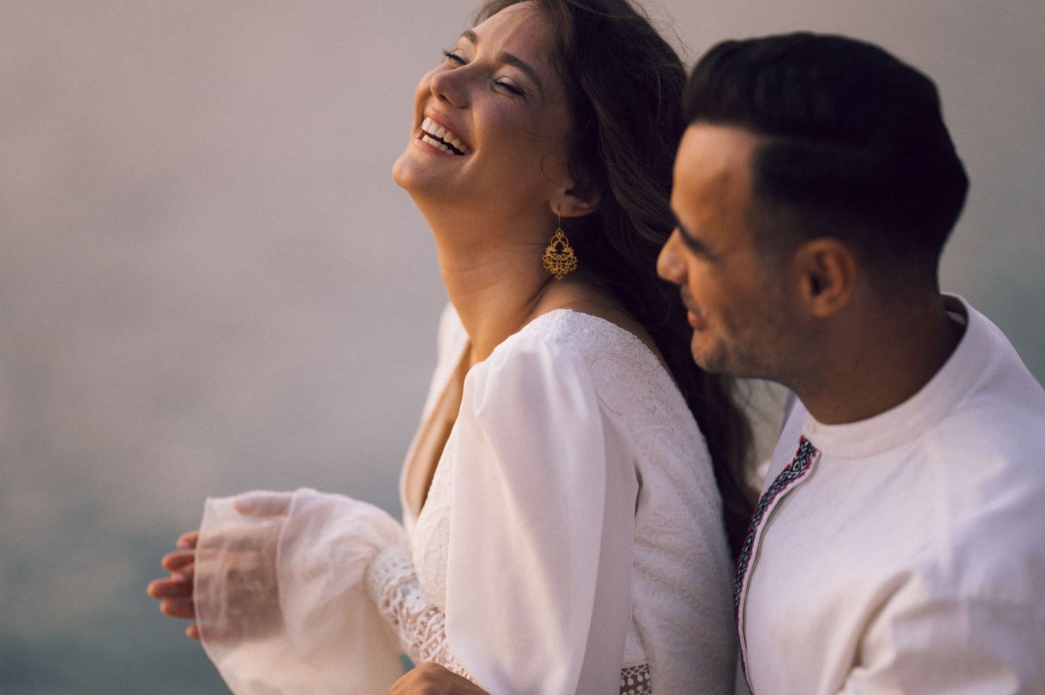 A joyful couple in white outfits laughing together outdoors, sharing a happy romantic moment.