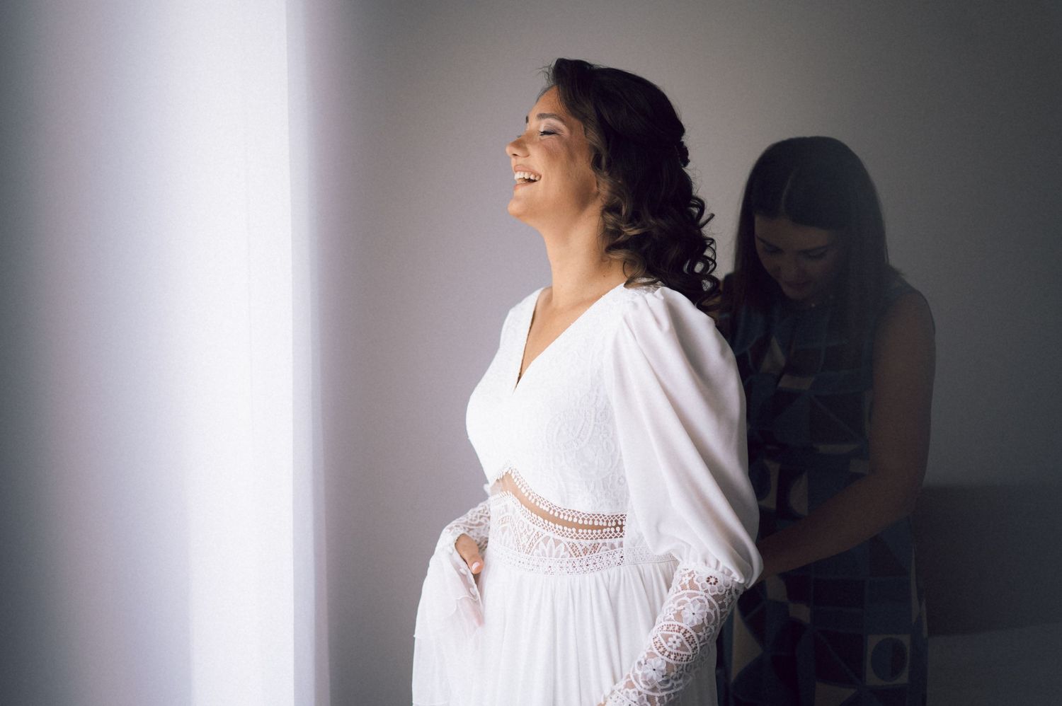 Smiling bride in white wedding dress being helped by bridesmaid near bright window.