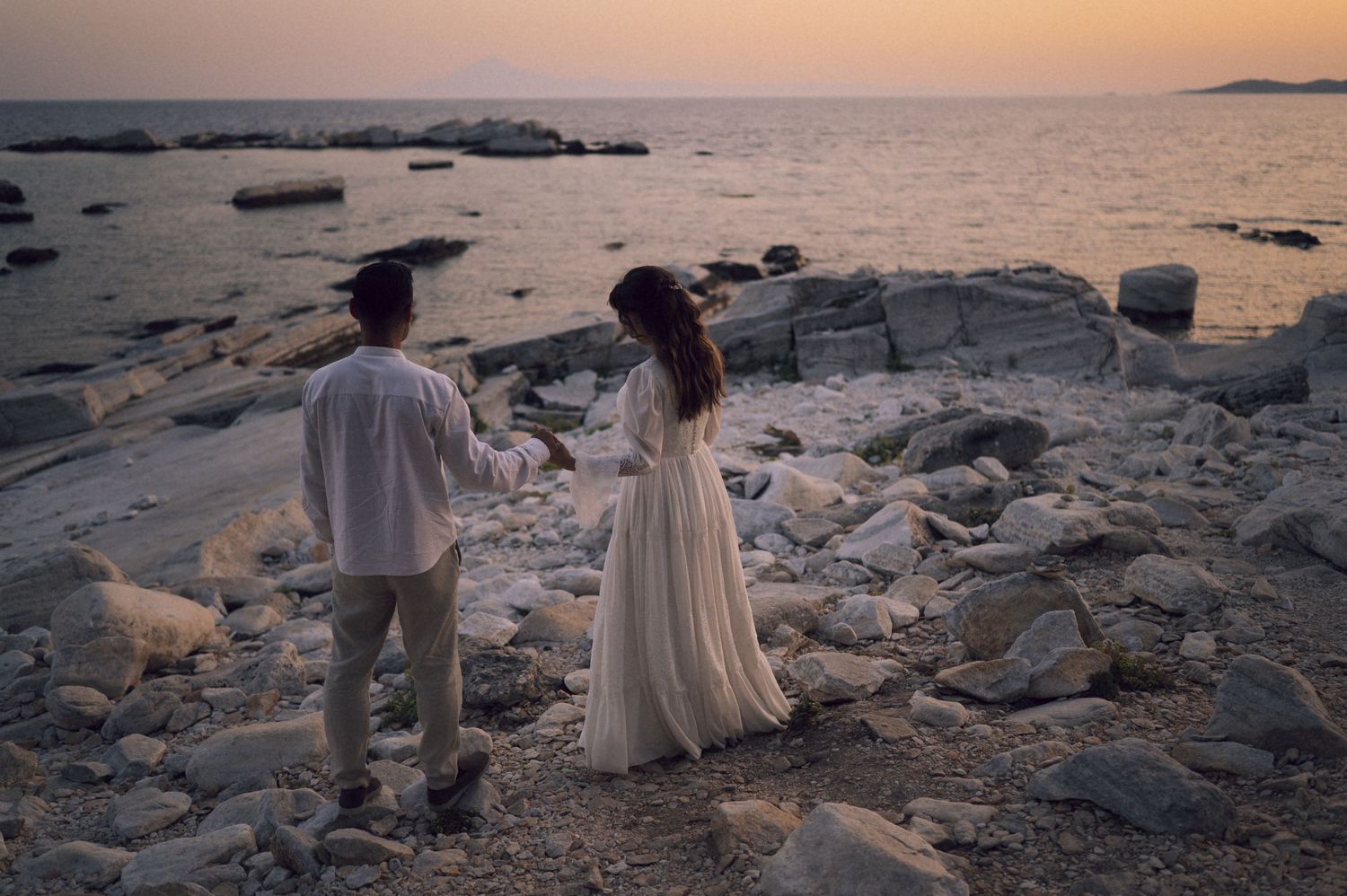 Couple holding hands on rocky beach at sunset, woman in white dress, romantic seaside photography.