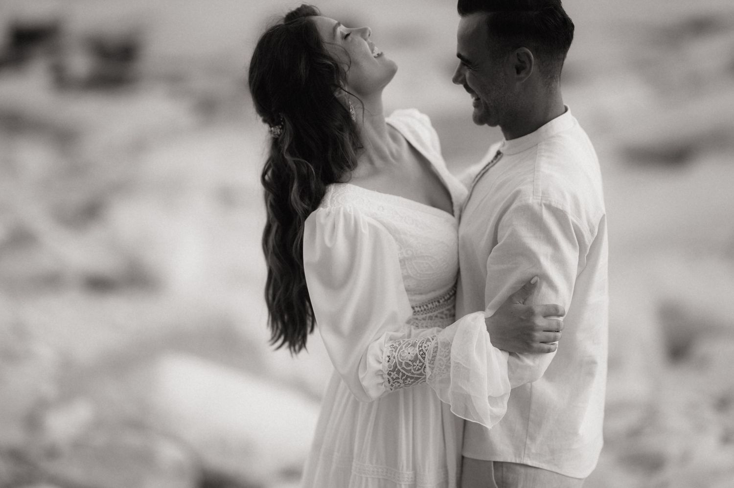 Black and white photo of a joyful couple embracing outdoors, woman in white dress laughing, man in white shirt smiling.