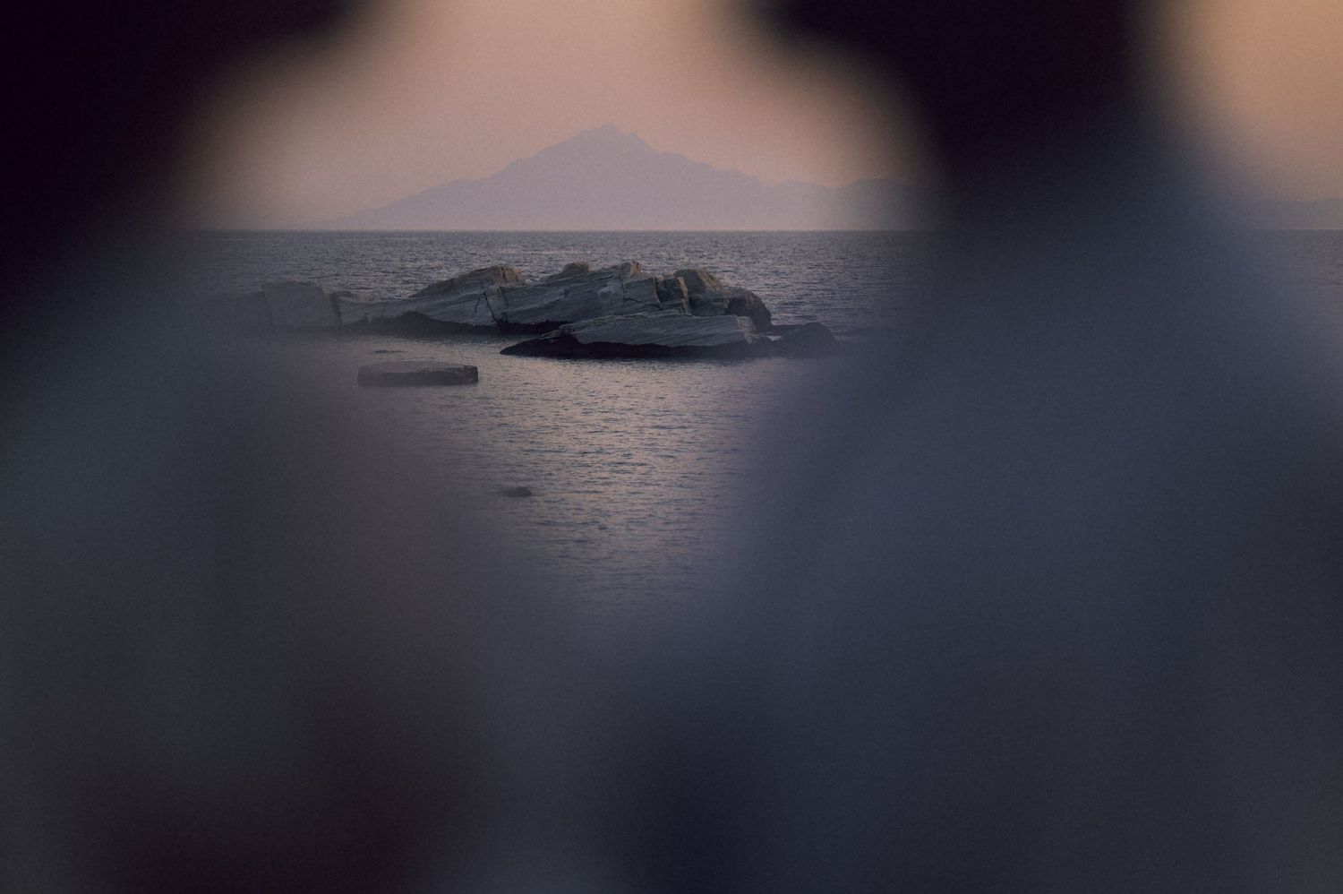 Misty seascape at dusk with rocky outcrop and distant mountain silhouette framed by blurred foreground.