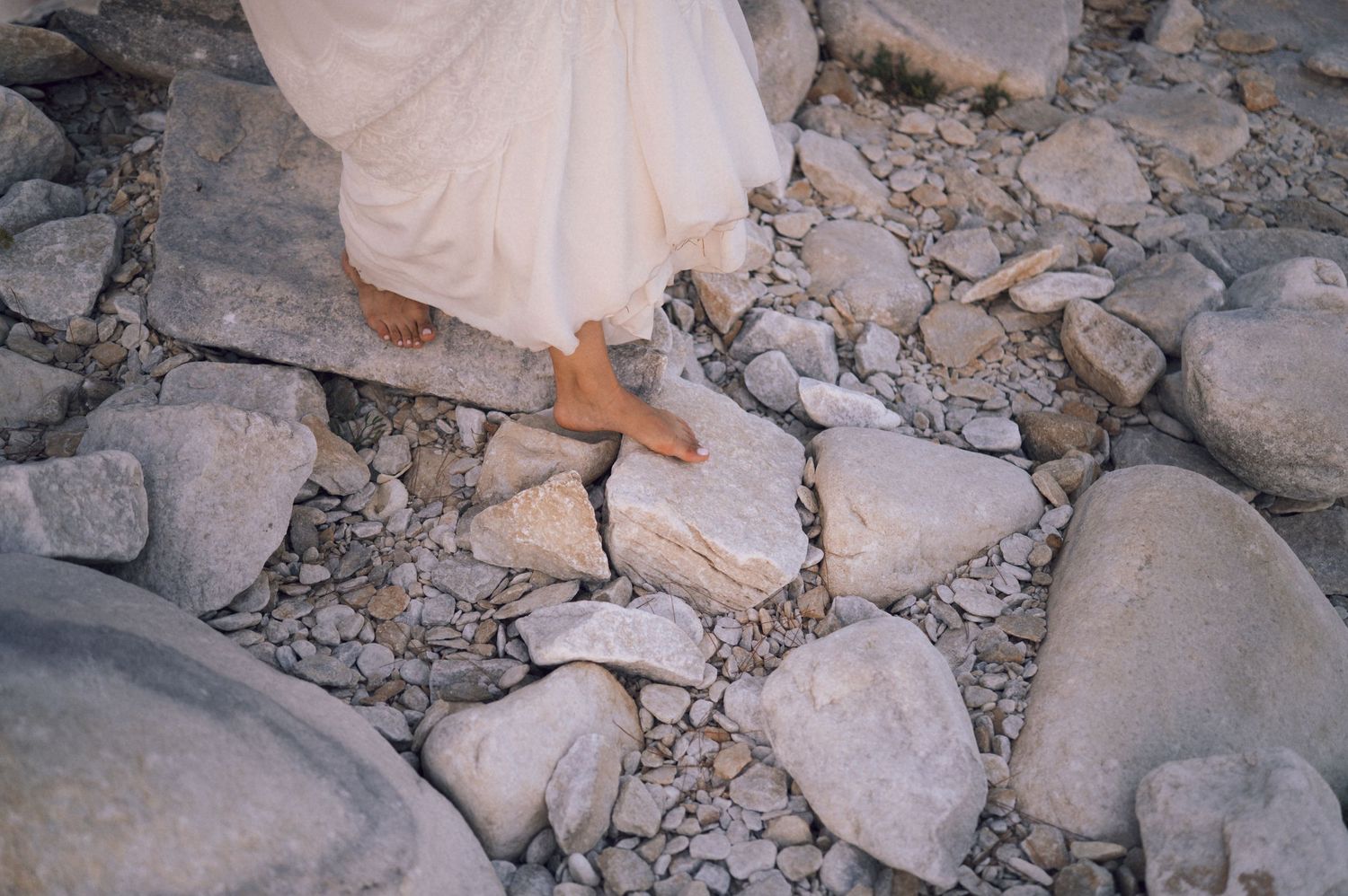 Barefoot person in white dress walking carefully over rocky terrain with large stones and pebbles.
