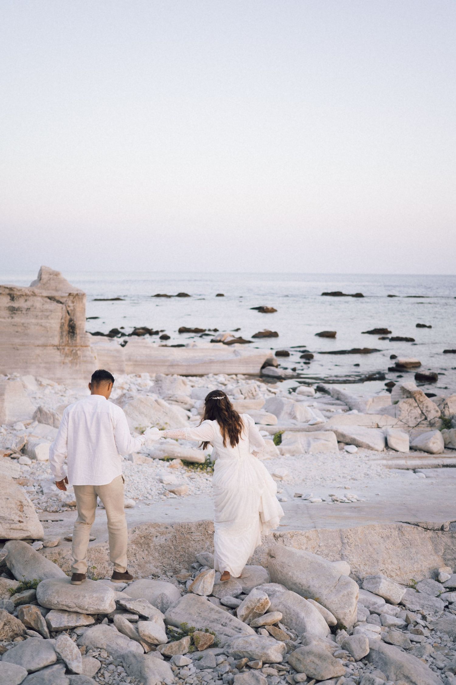 Couple in white attire walking hand-in-hand on rocky beach at sunset with calm ocean in background.