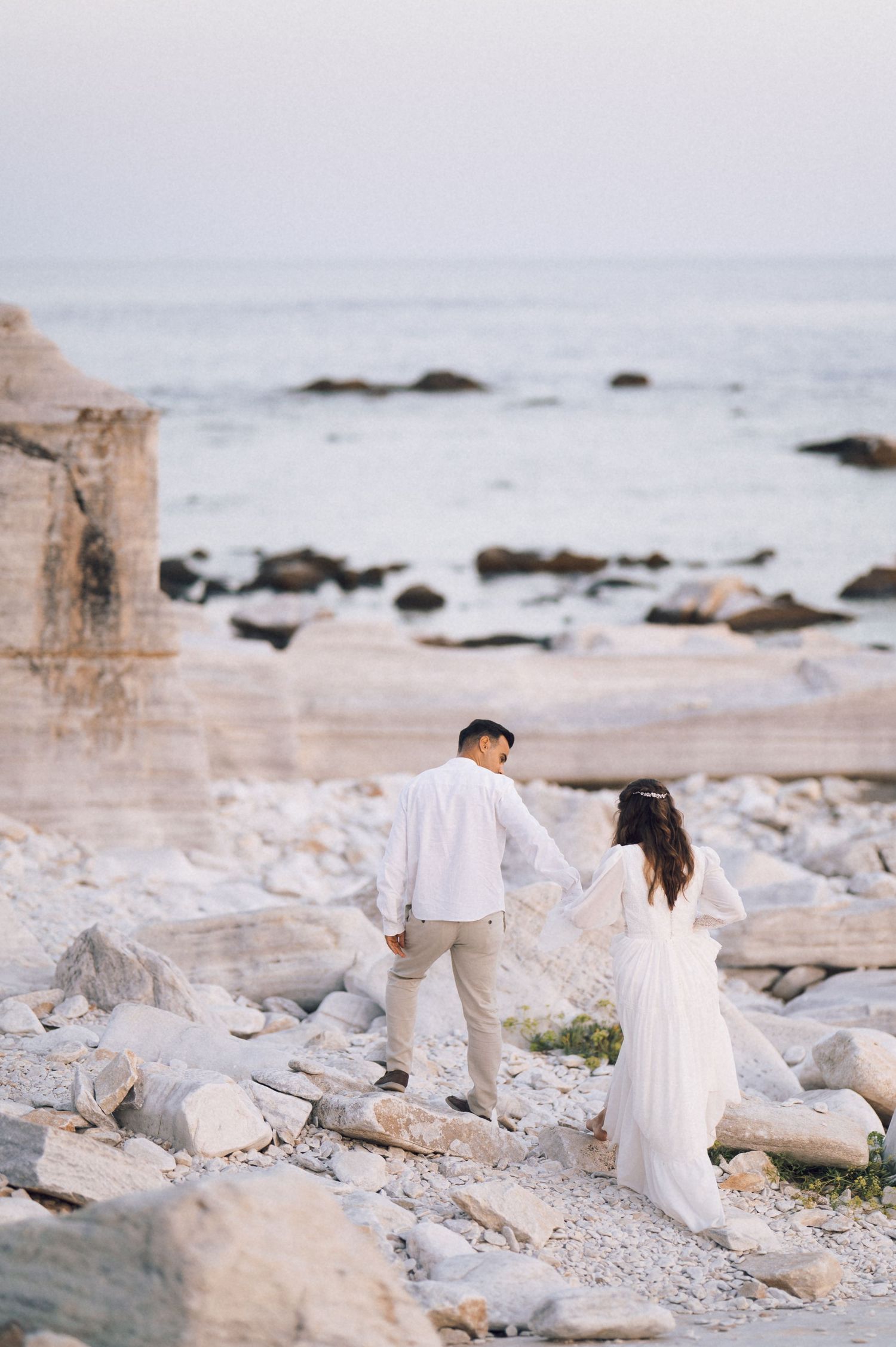 Couple in white outfits holding hands on rocky beach shore near ocean ruins at dusk.