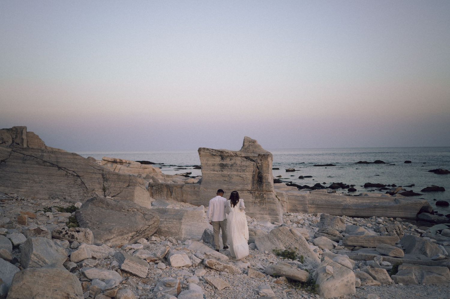Couple standing among ancient coastal ruins at dusk with rocky shoreline and pastel sky backdrop.