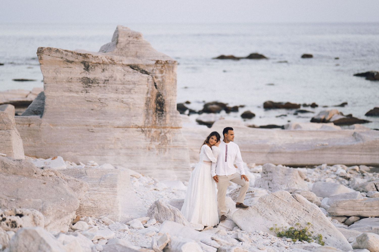 Couple in white wedding attire posing on rocky coastal cliffs with turquoise ocean waves in the background.