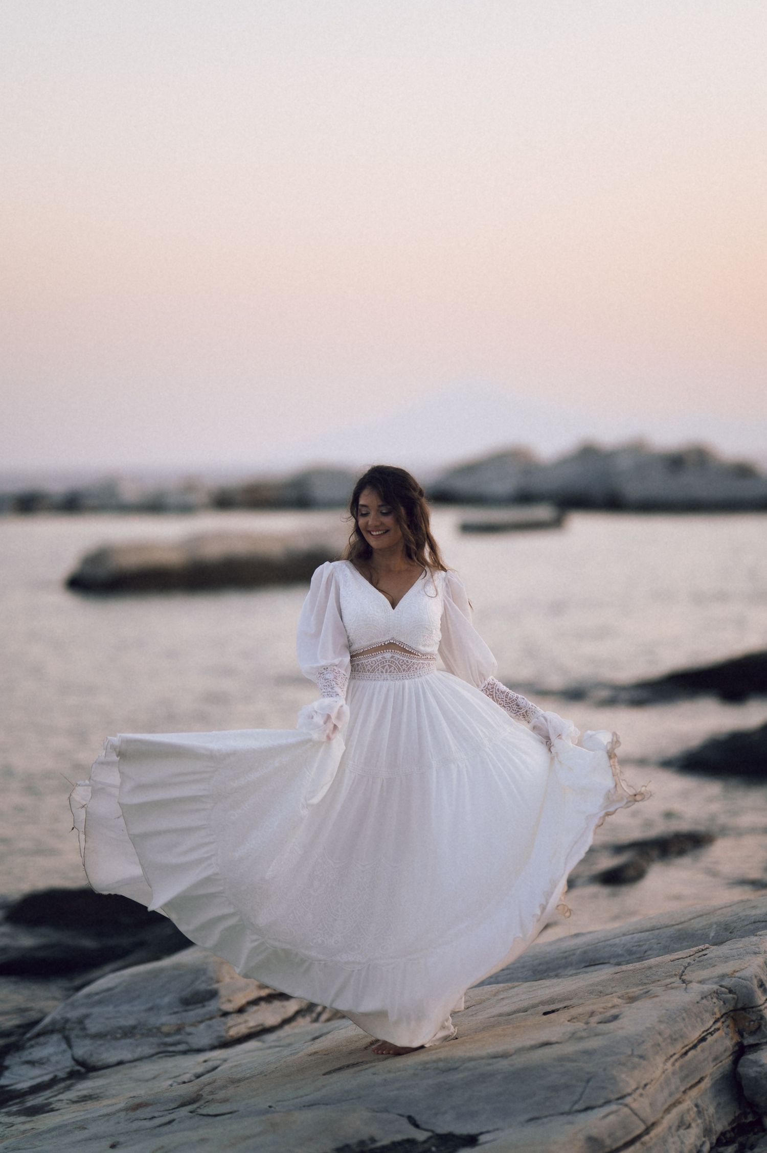 Woman in flowing white dress standing on rocky shore at sunset by the sea.