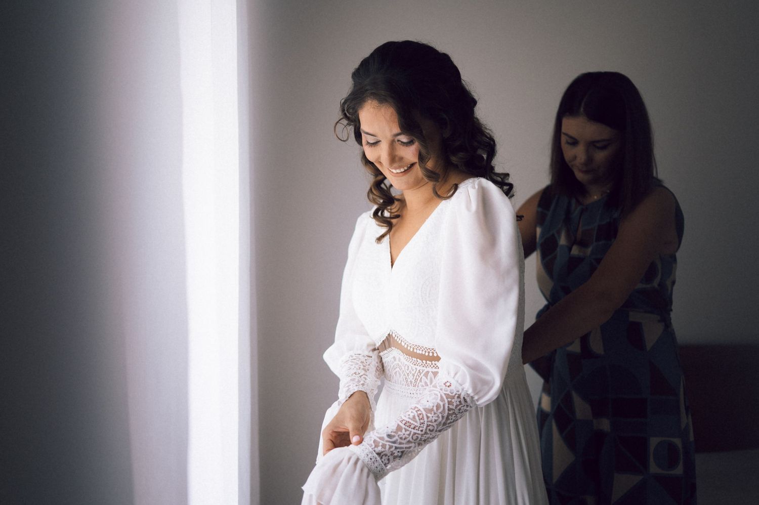 Bride smiling in white wedding dress being helped by bridesmaid near bright window.