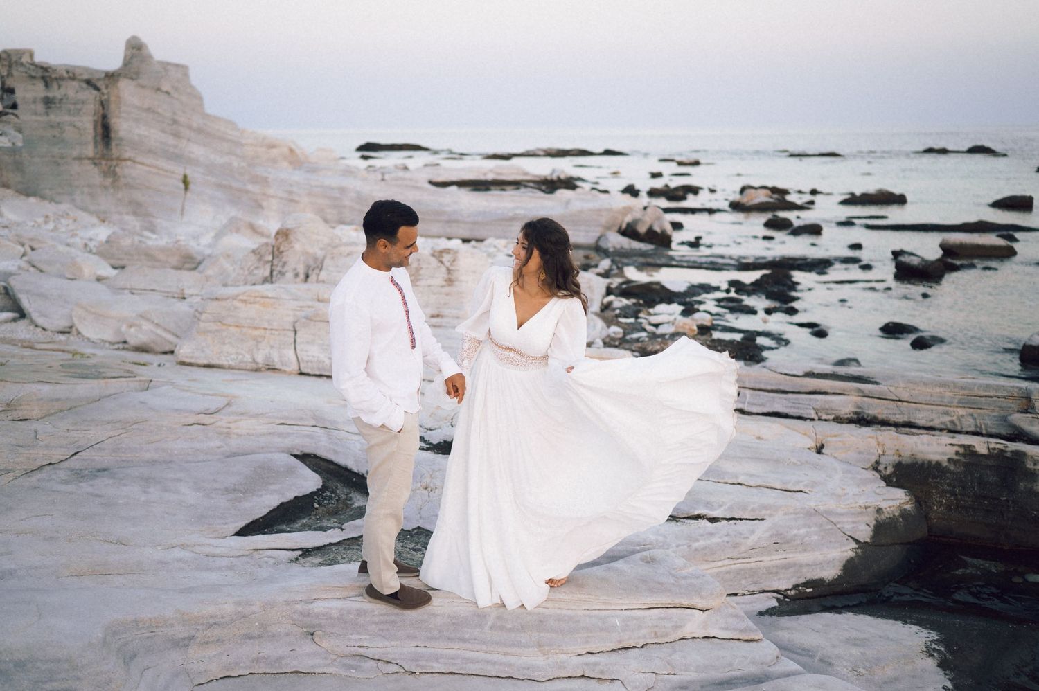 Couple in white attire holding hands on rocky coastal cliffs with calm sea backdrop at dusk.