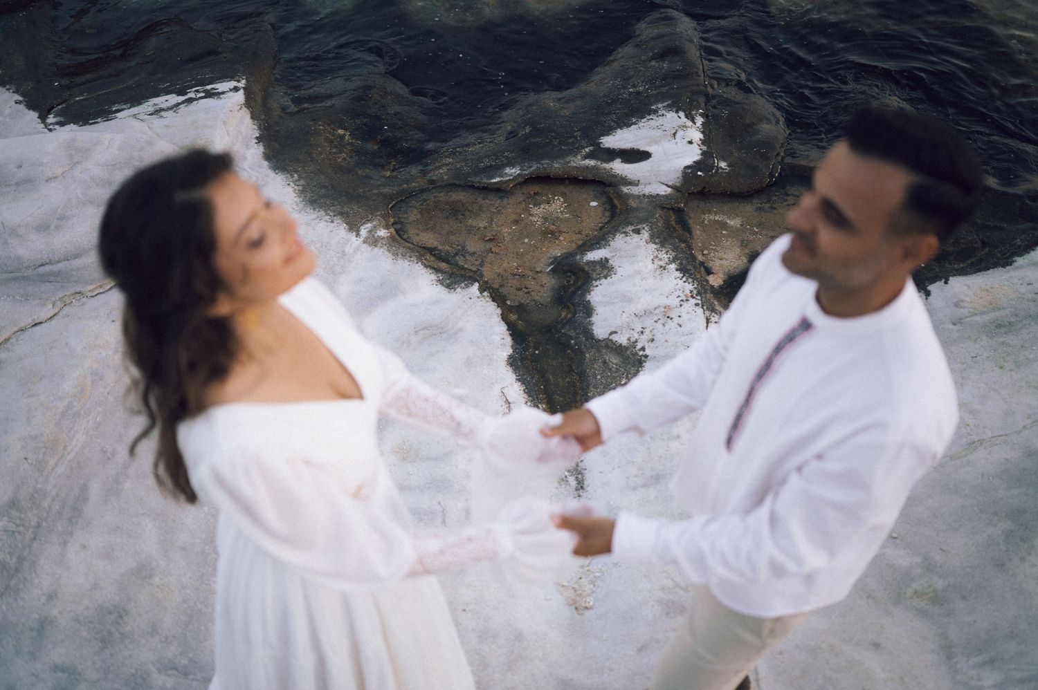 Couple in white outfits holding hands dancing on rocky coastal background during romantic photoshoot.
