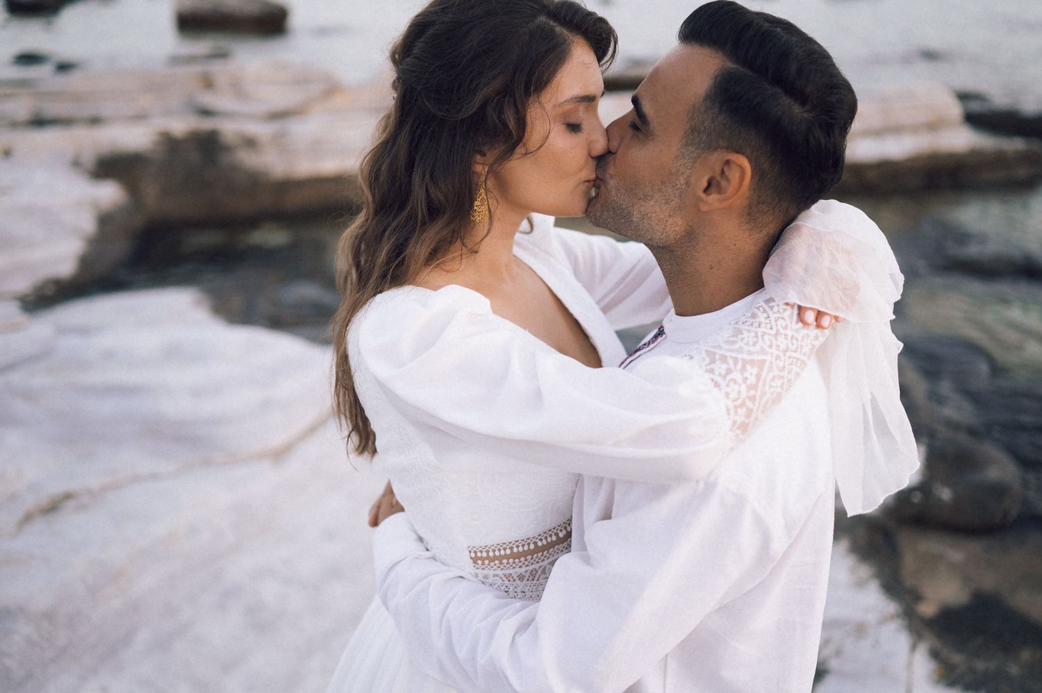 Romantic couple sharing a kiss on rocky beach, both wearing white outfits, with ocean backdrop.