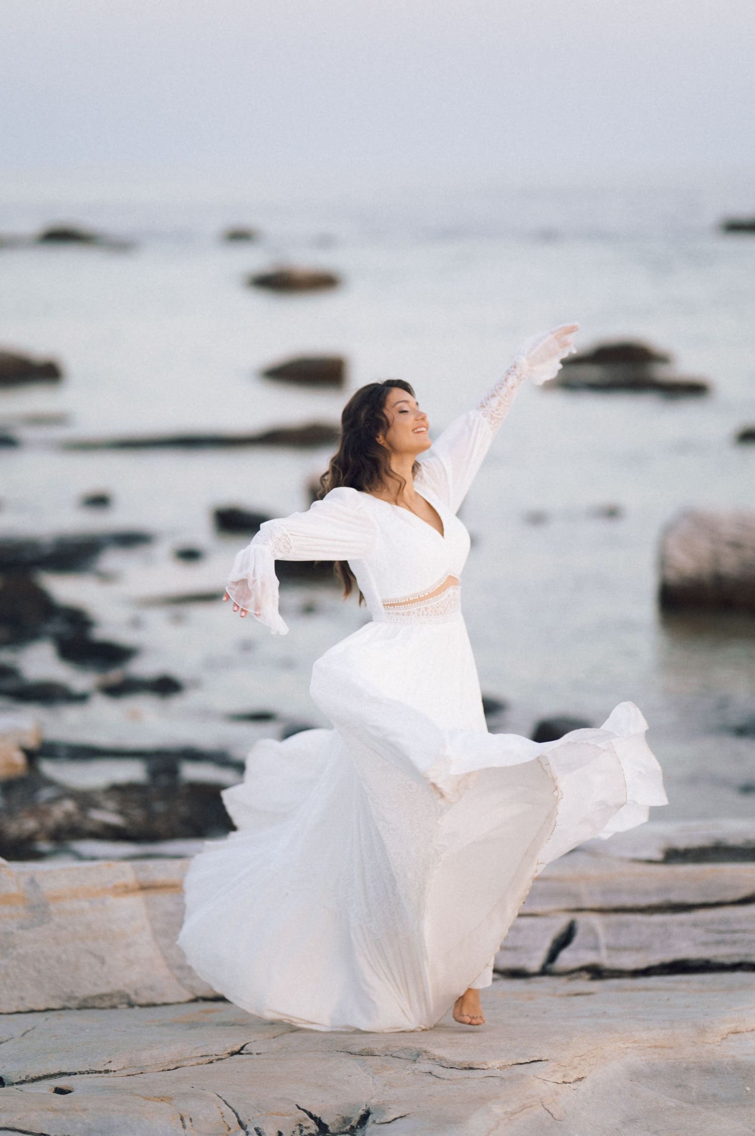 Woman in flowing white dress dancing gracefully on rocky shoreline with arms outstretched by the sea.