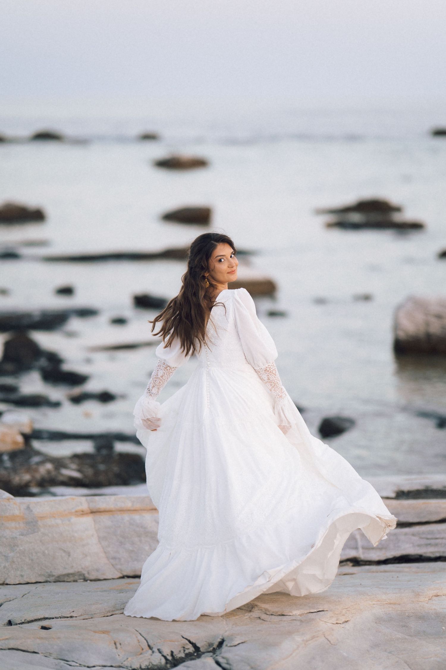 A woman in a flowing white lace wedding dress stands on rocky shore with ocean backdrop.