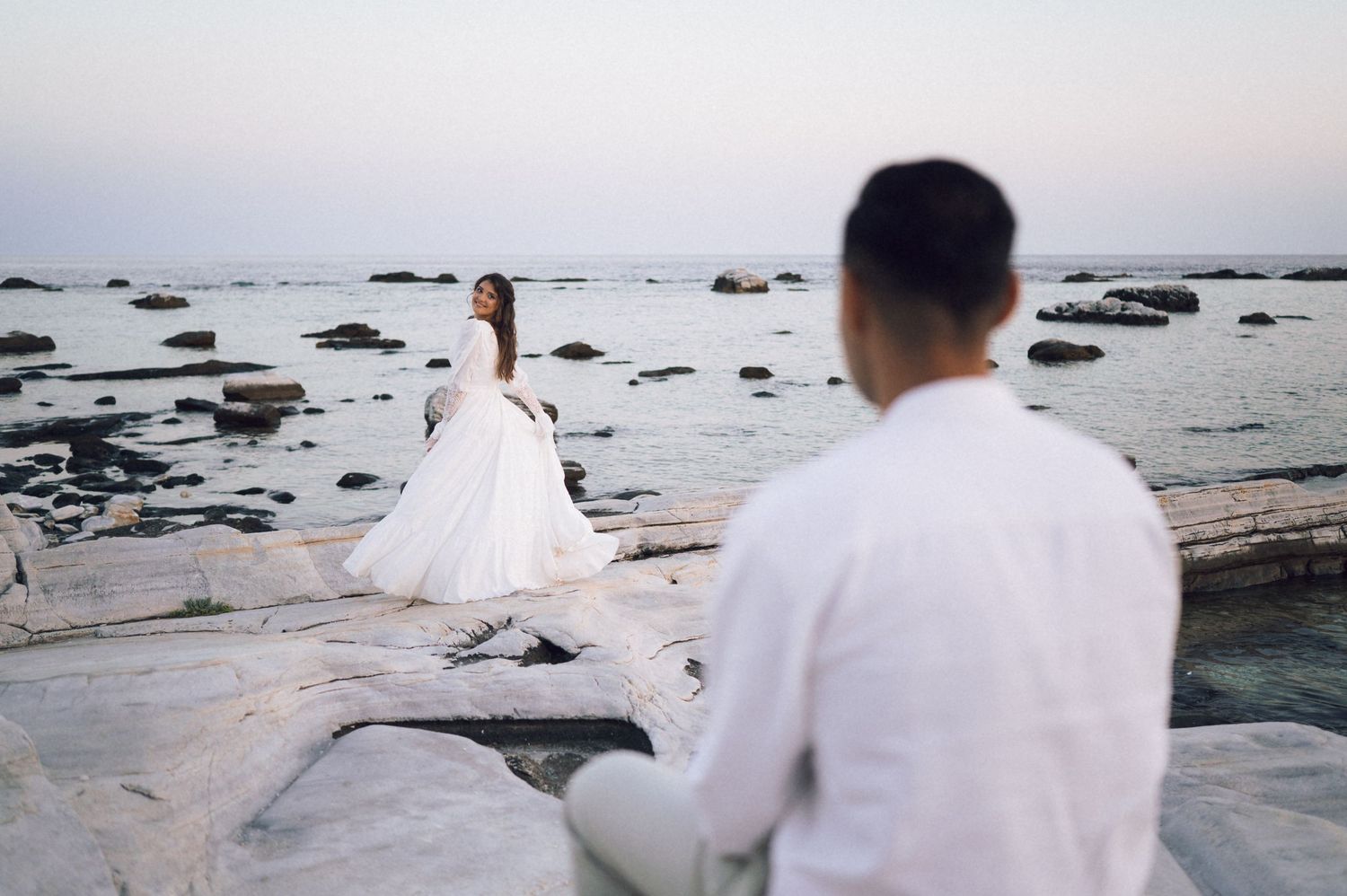 Bride in white gown stands on rocky seaside shore as groom in white shirt watches during romantic wedding photo session.