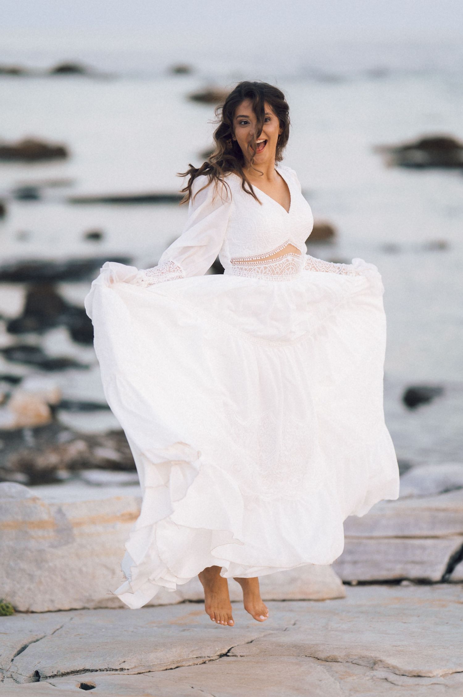 A joyful bride in a flowing white boho wedding dress jumps barefoot on coastal rocks by the sea.