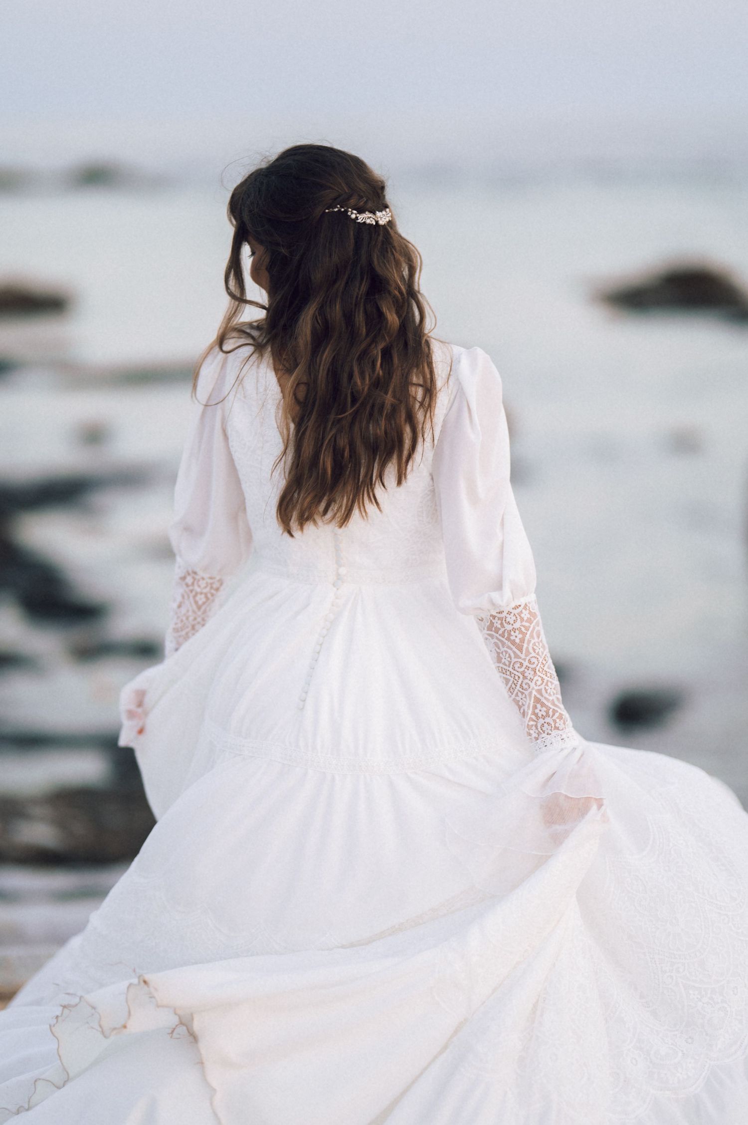 Rear view of a bride in a white boho gown with lace sleeves and floral hair crown by the ocean.