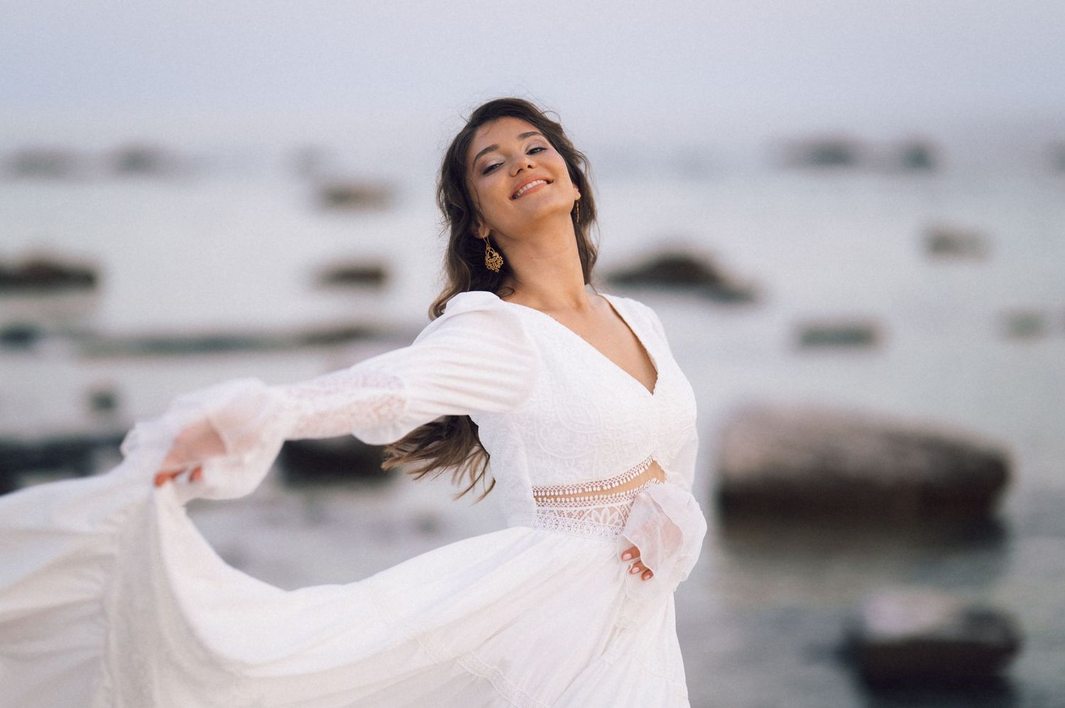 Woman in flowing white dress dancing gracefully by the ocean with rocks in the background.