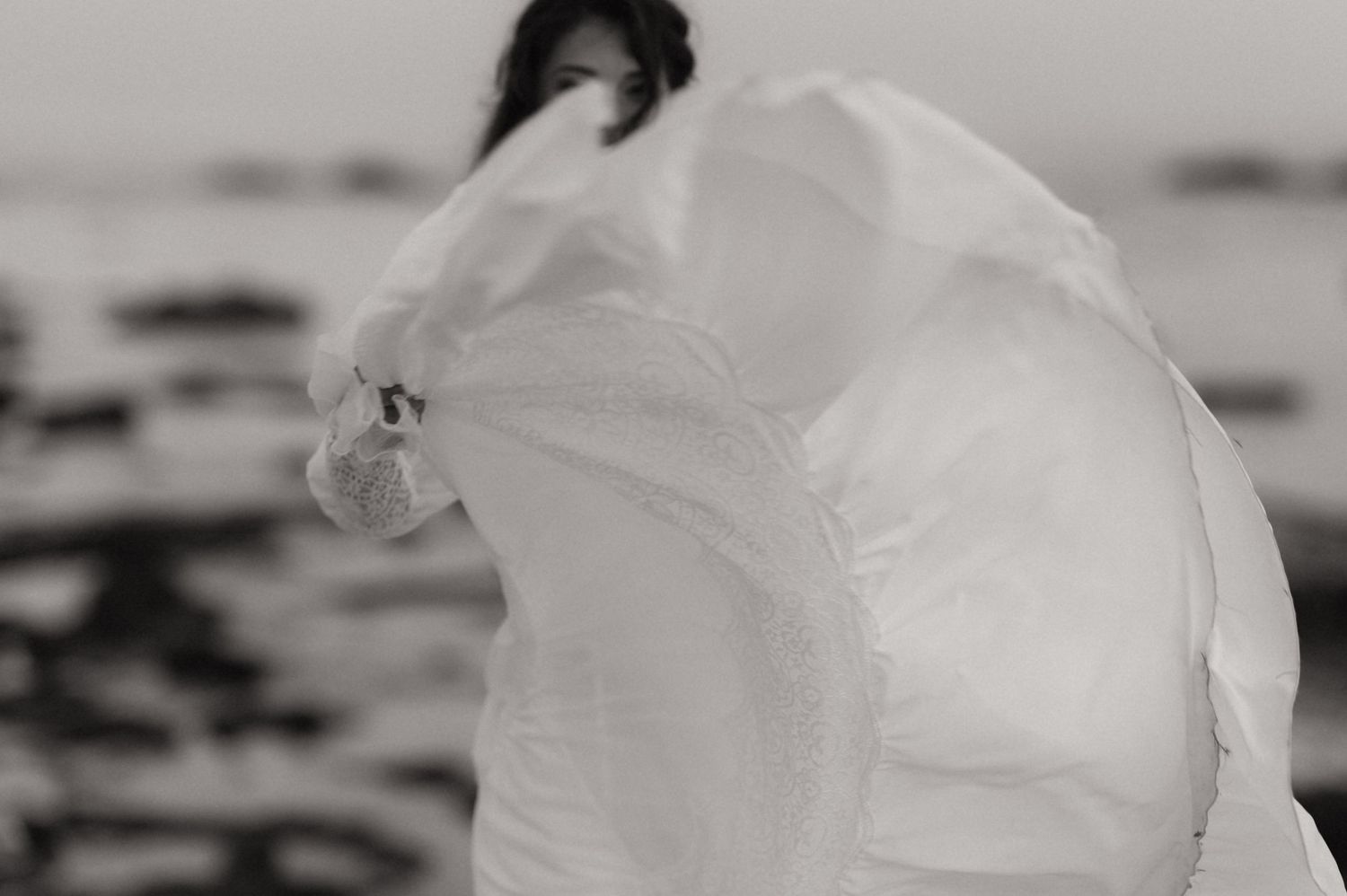 Black and white photo of a woman in a flowing white dress billowing in the wind by the ocean shore.