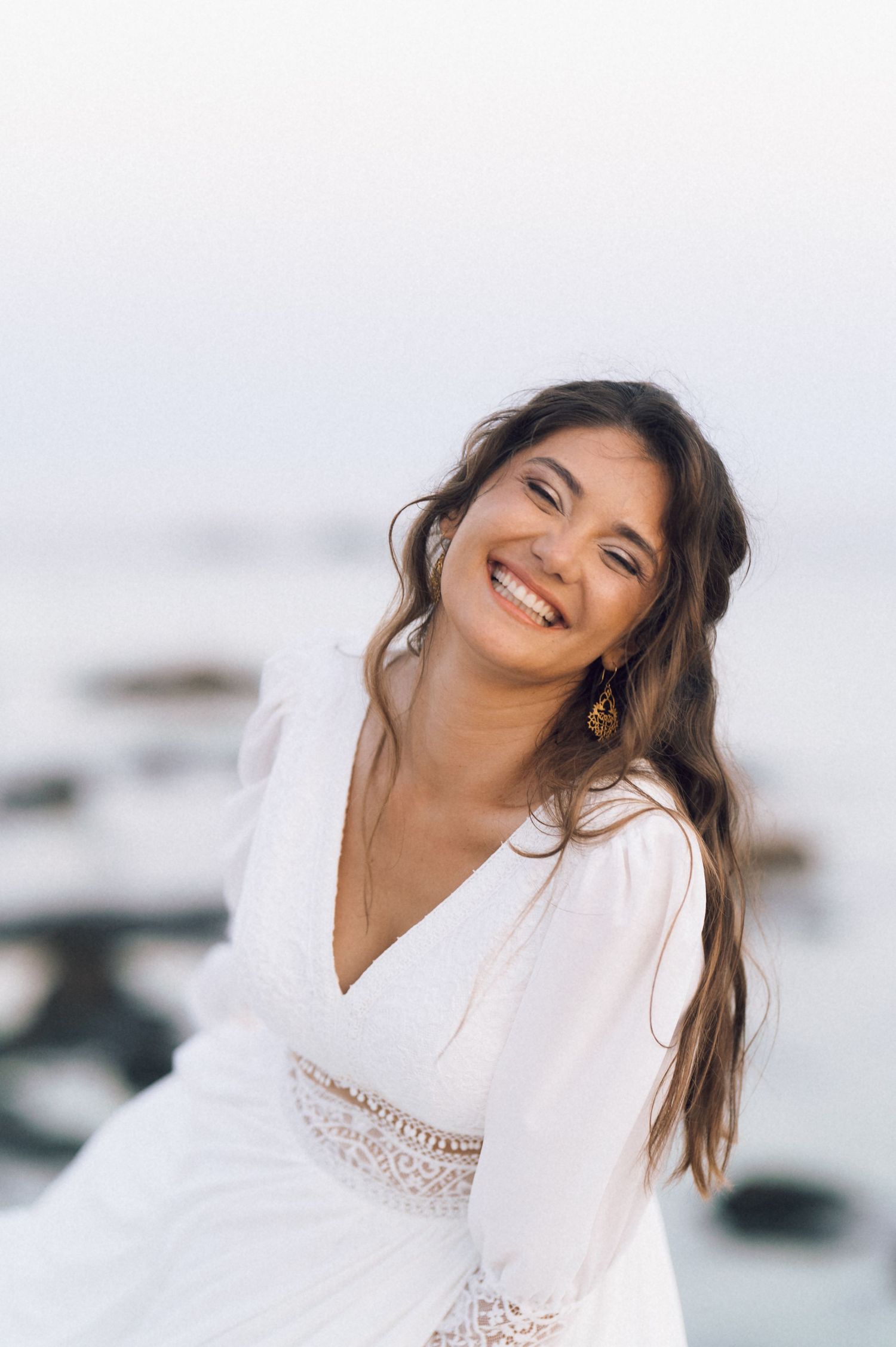 Smiling young woman with long brown hair wearing a white lace dress and gold earrings posing near the ocean.