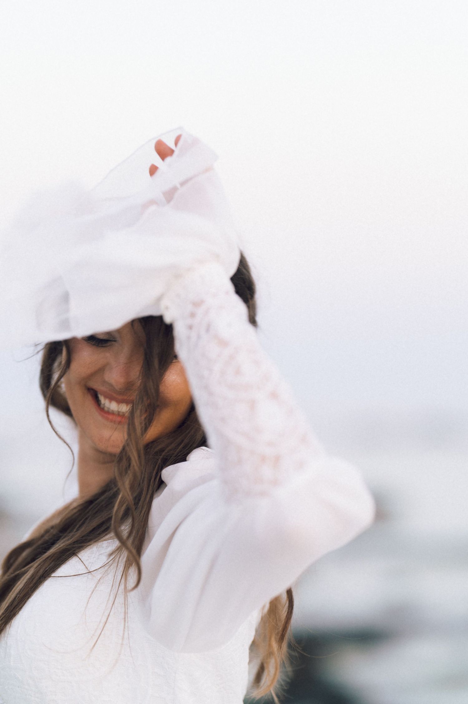 Smiling bride in white lace gloves and veil laughing outdoors on a breezy day.