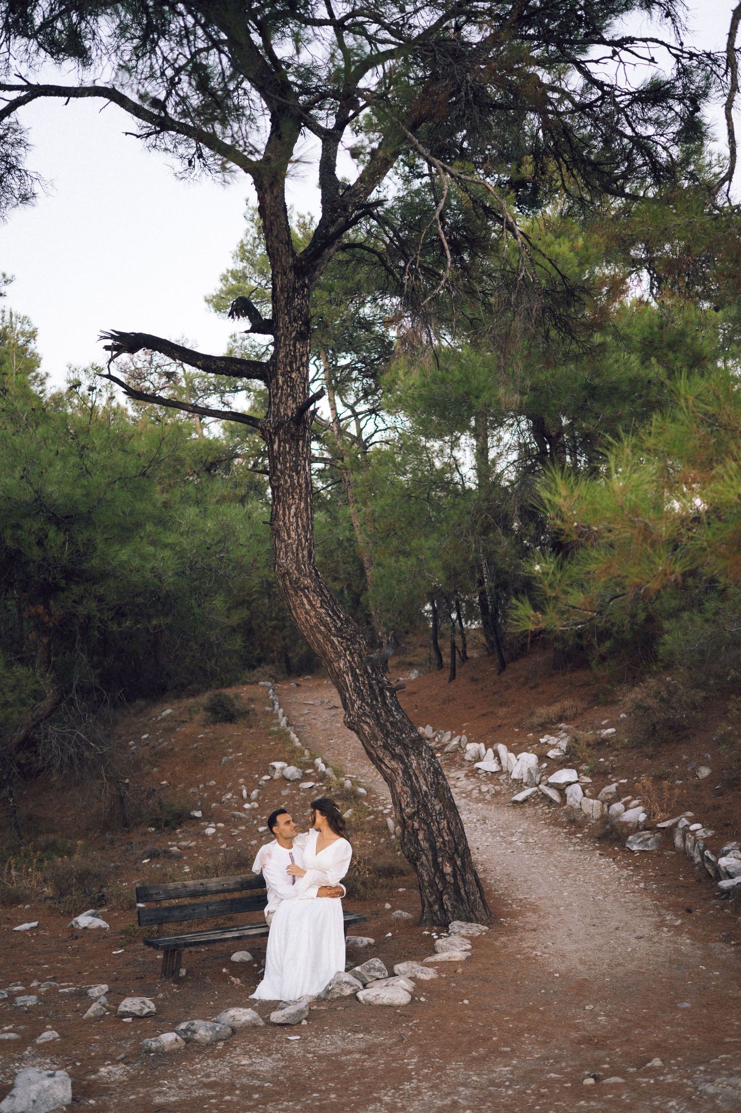 Couple in white robes embracing under a pine tree on a rocky forest path with a wooden bench nearby.