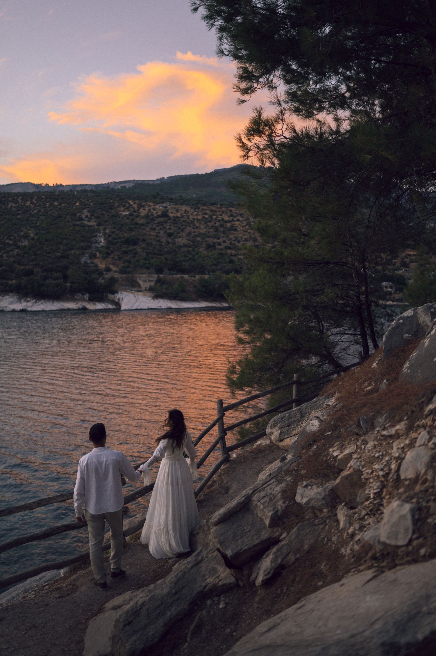 Couple in wedding attire holding hands on rocky shore at sunset by a serene lake with lush green hills.