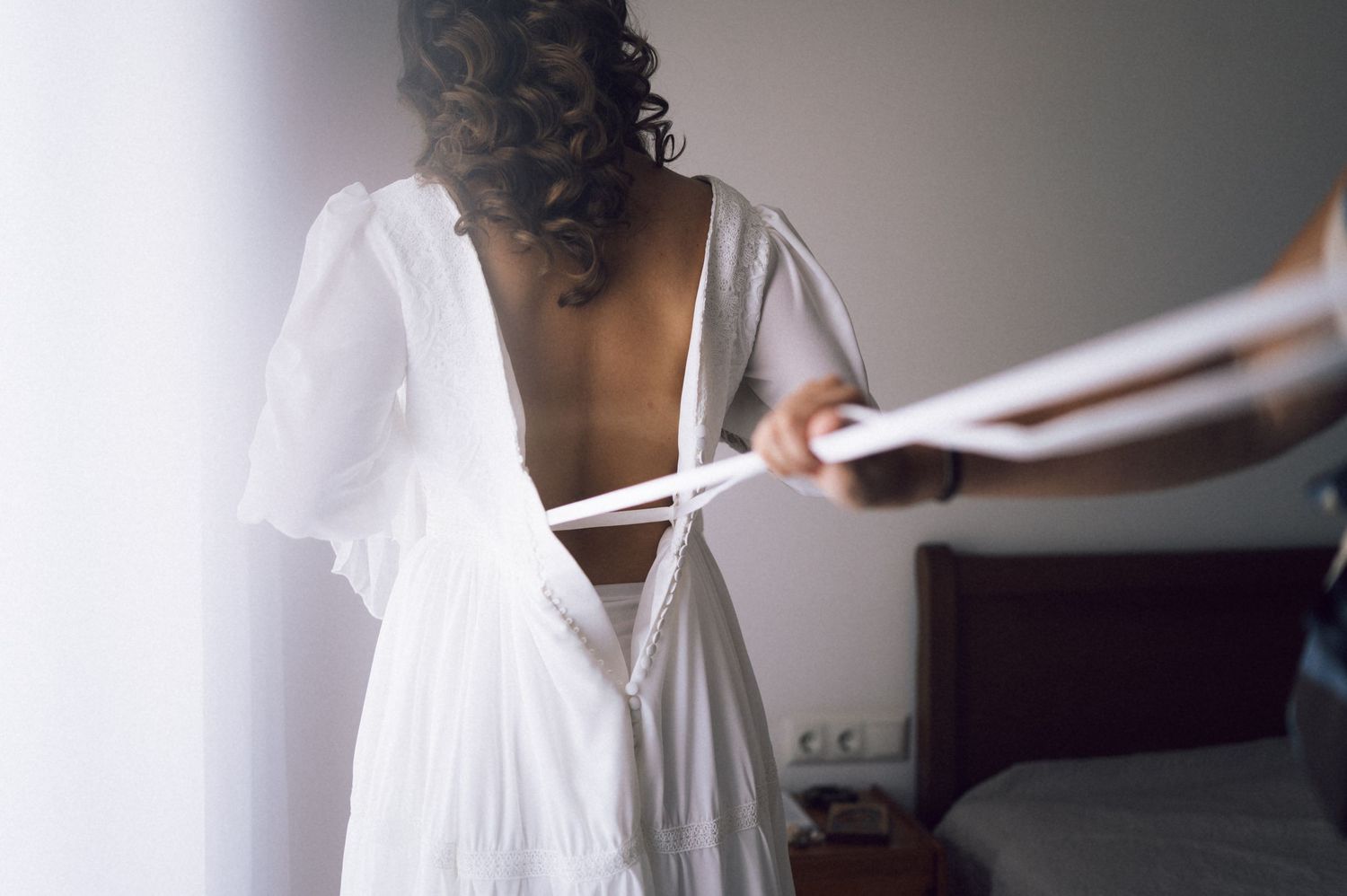 Bride in white open-back wedding dress being laced up by assistant in bright bedroom.