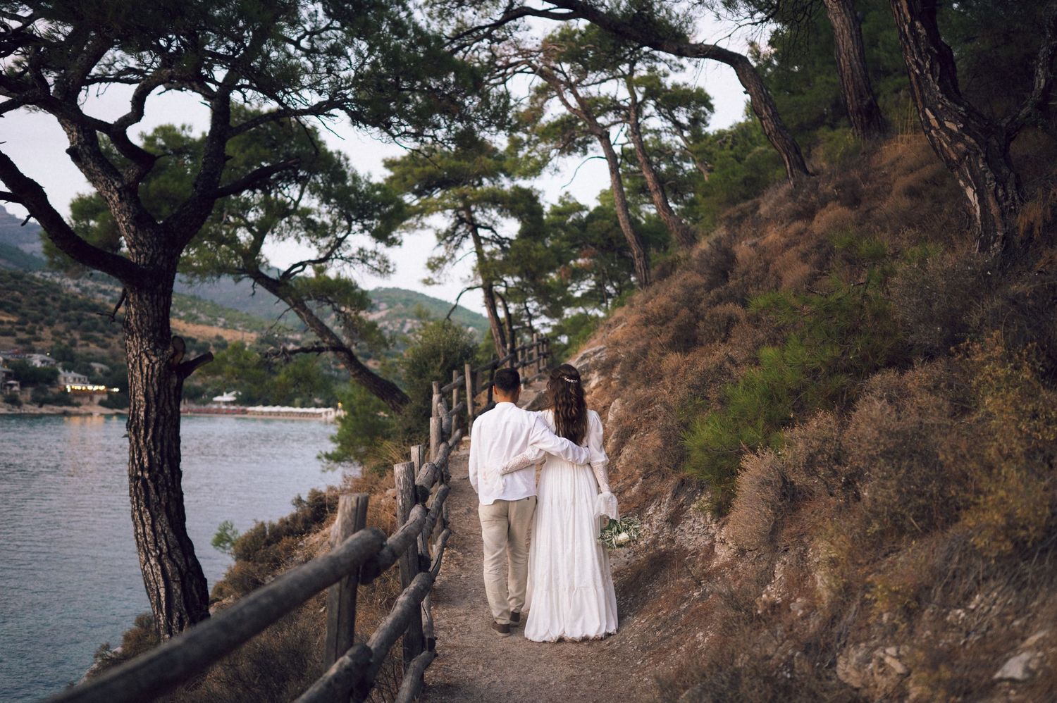 Couple in wedding attire walking along a scenic coastal path surrounded by pine trees and hillside views.