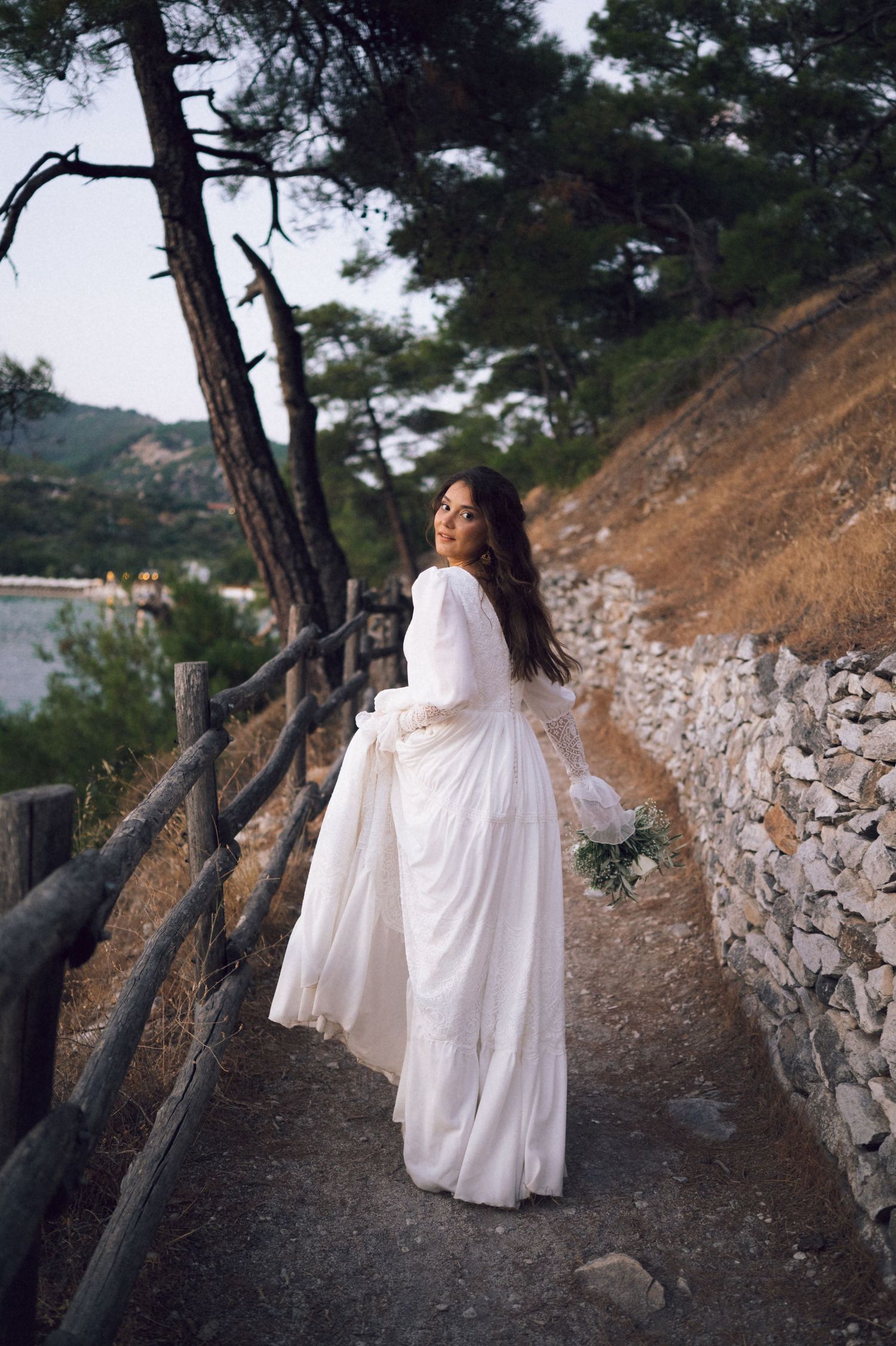 Bride in white gown walks scenic coastal path with wooden fence, holding bouquet, surrounded by pine trees.