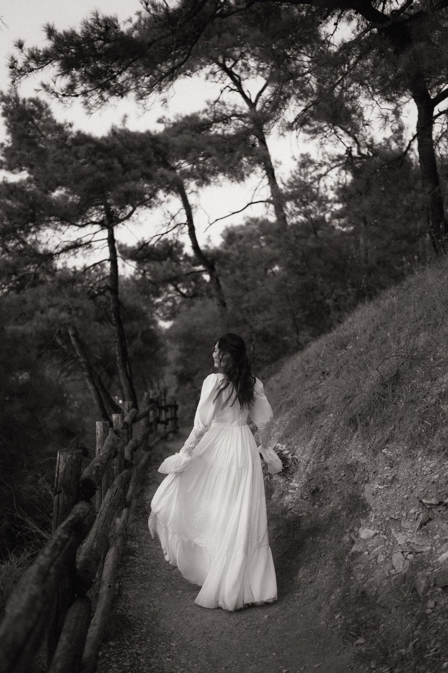Black and white photo of a bride in a flowing white gown walking along a forest path holding a bouquet.