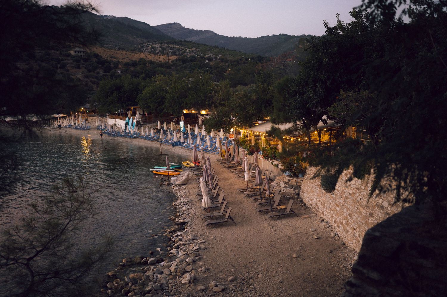 Twilight beach resort with lounge chairs and umbrellas along a calm cove, surrounded by lush green hills.