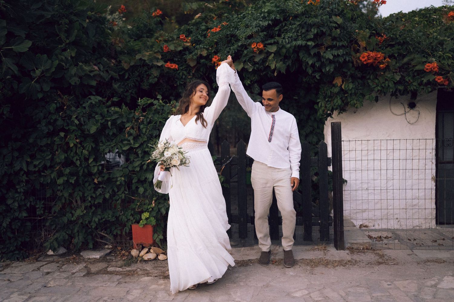 Bride and groom dancing outdoors in front of lush green foliage with orange flowers on their wedding day.
