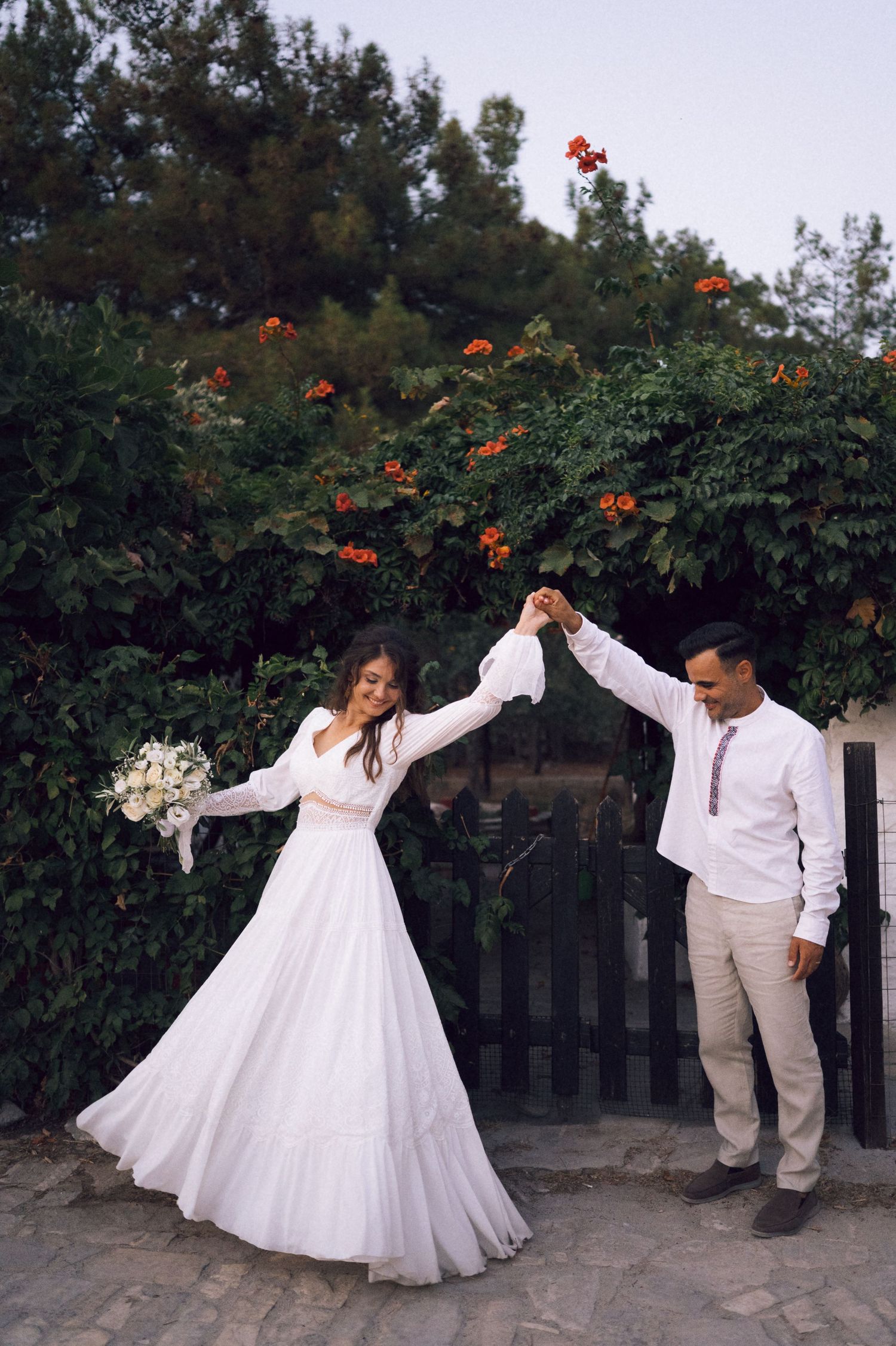 A bride in a white gown and groom in embroidered shirt dance joyfully outdoors amid lush green trees with red flowers.