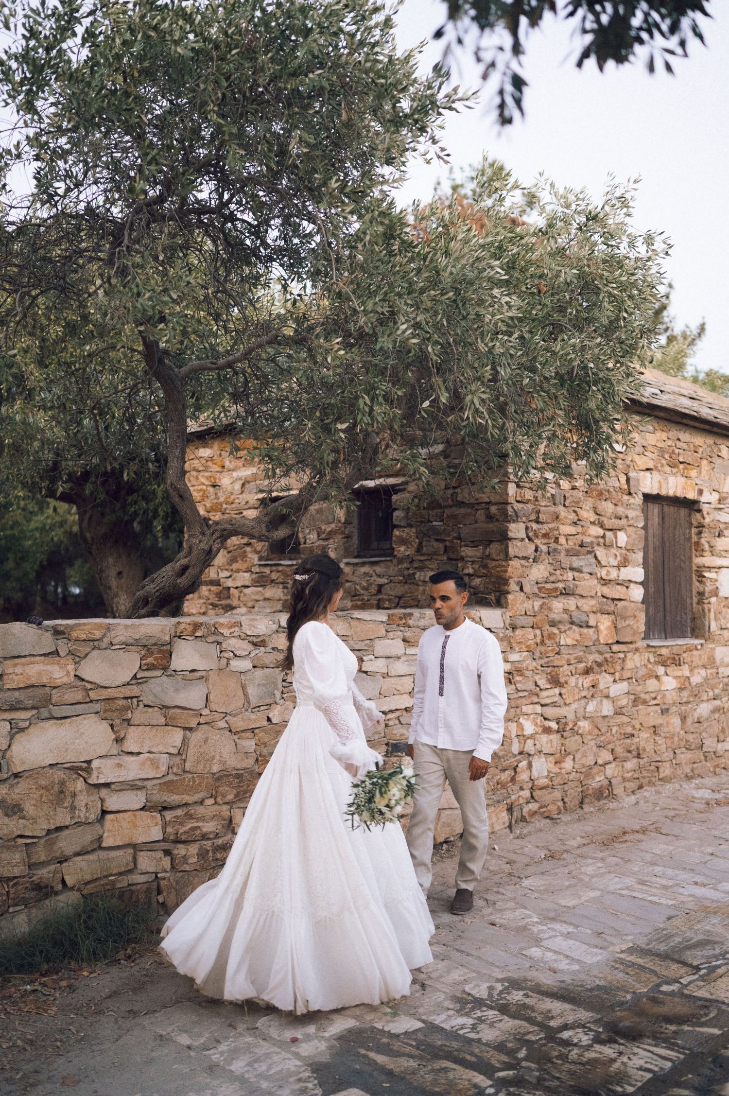 Bride and groom walk hand in hand beside a rustic stone wall with an olive tree backdrop.